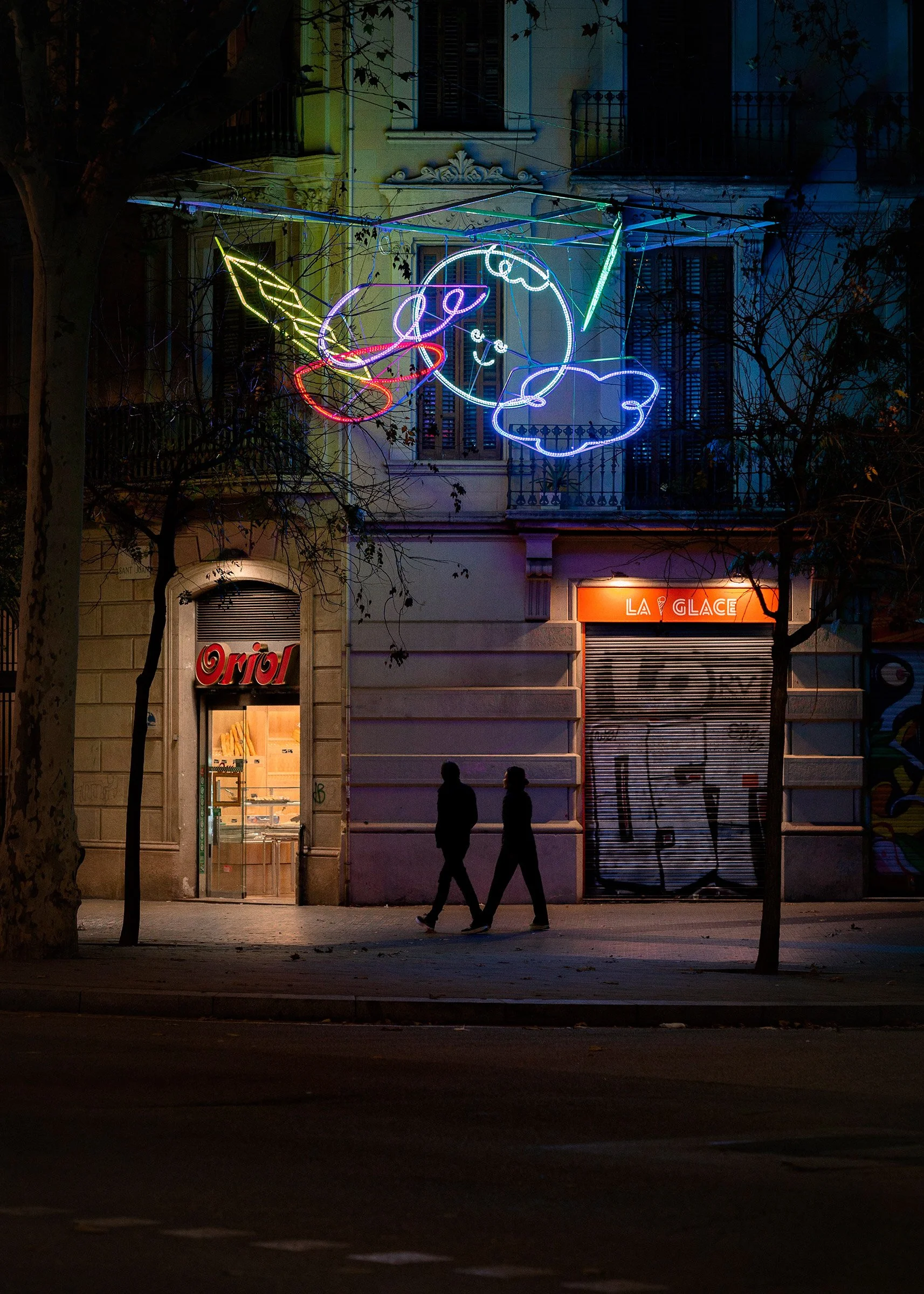 Side-angle night photo of an illuminated cloud cluster of festive icons along the Barcelona pavement.