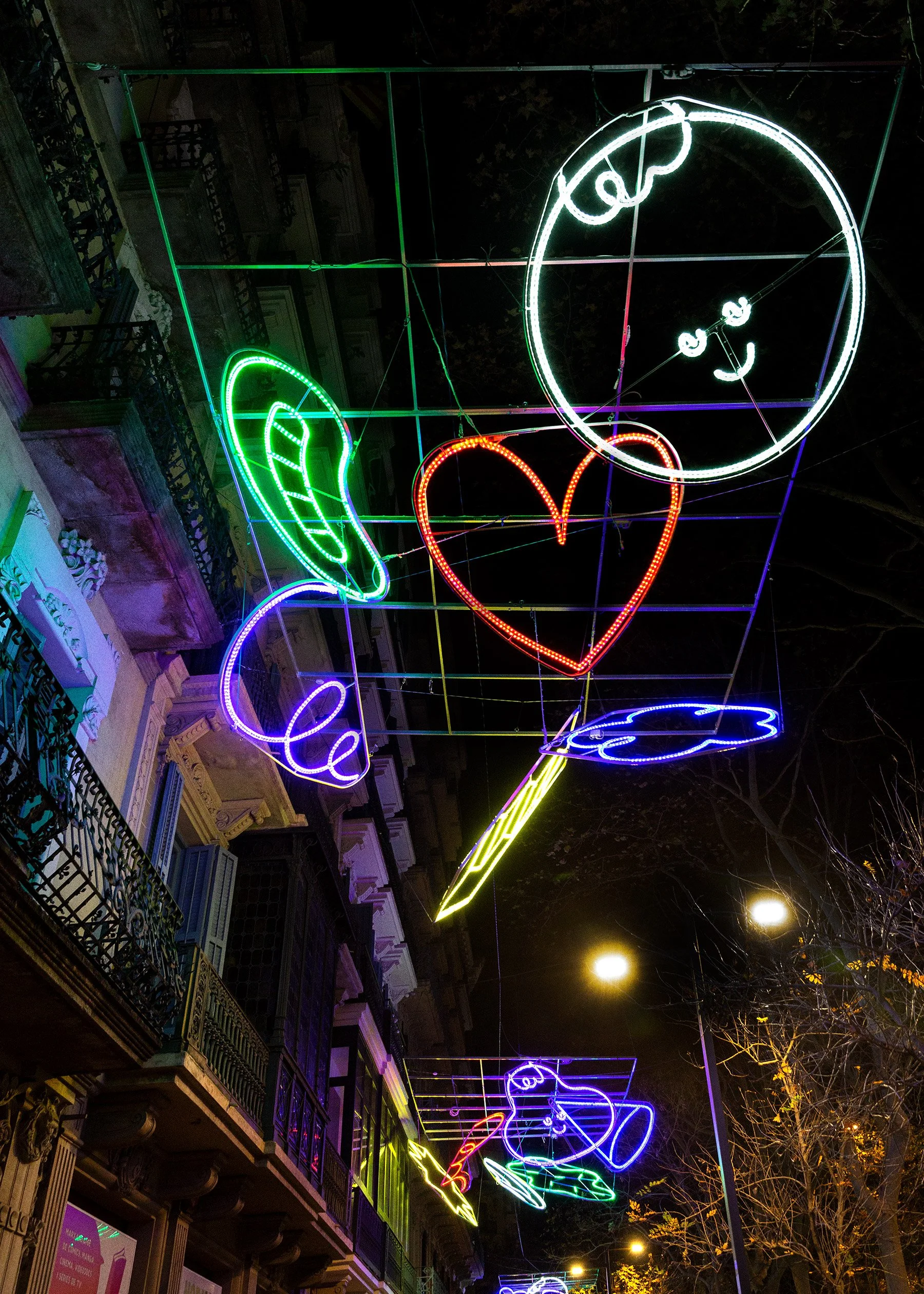 Low-angle night photo of colorful red, green, and purple illuminated Christmas icons hanging above the street.
