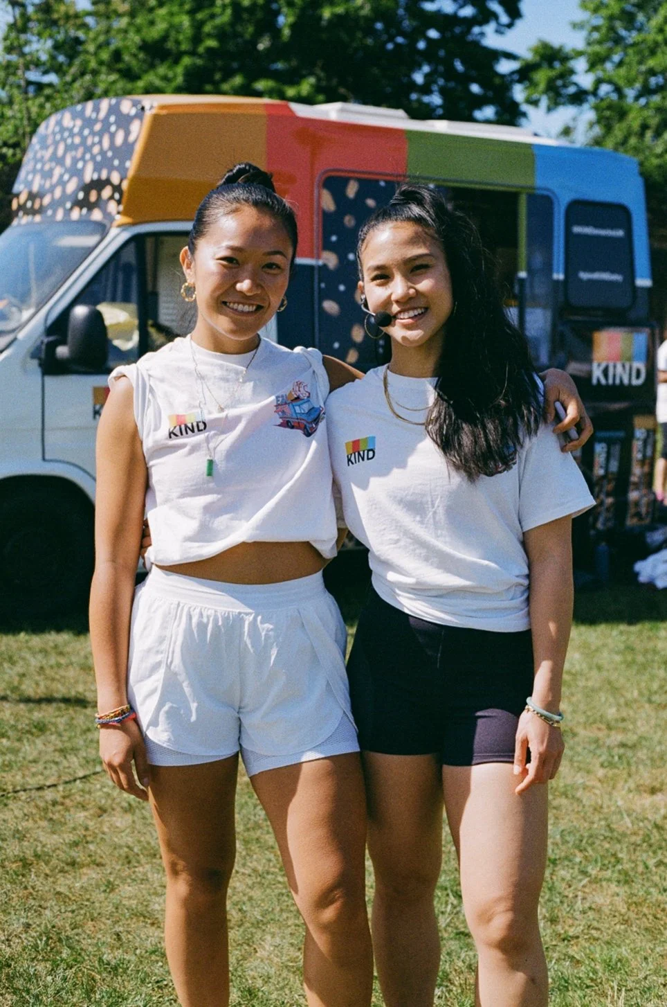 Two young women smiling, standing outdoors with their arms around each other, in front of a colorful food truck. They are wearing white t-shirts with a rainbow-colored 'KIND' logo.