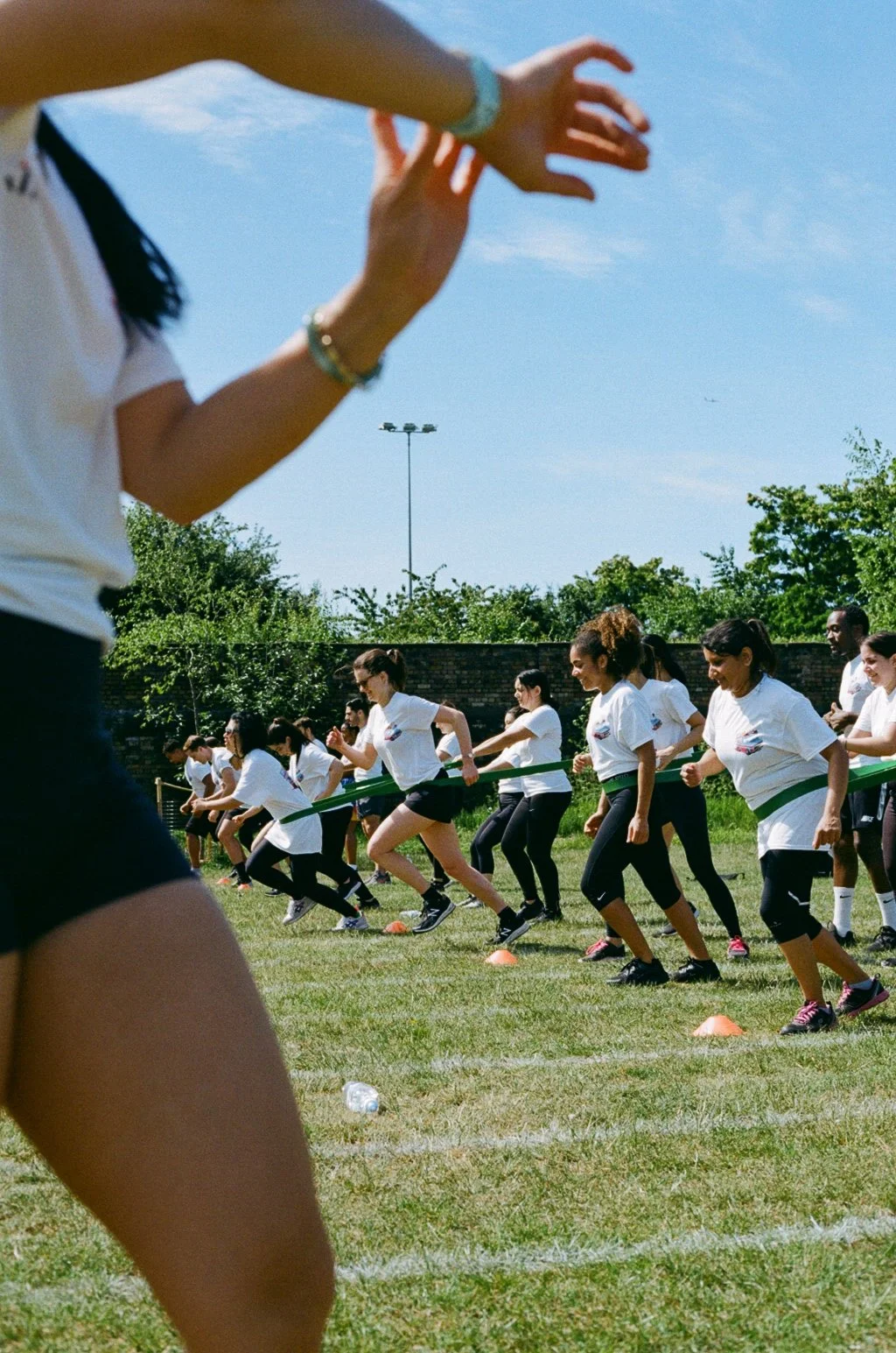 A group of women participating in a relay race or outdoor team activity, running across a grassy field with some holding a green baton, under a clear blue sky.