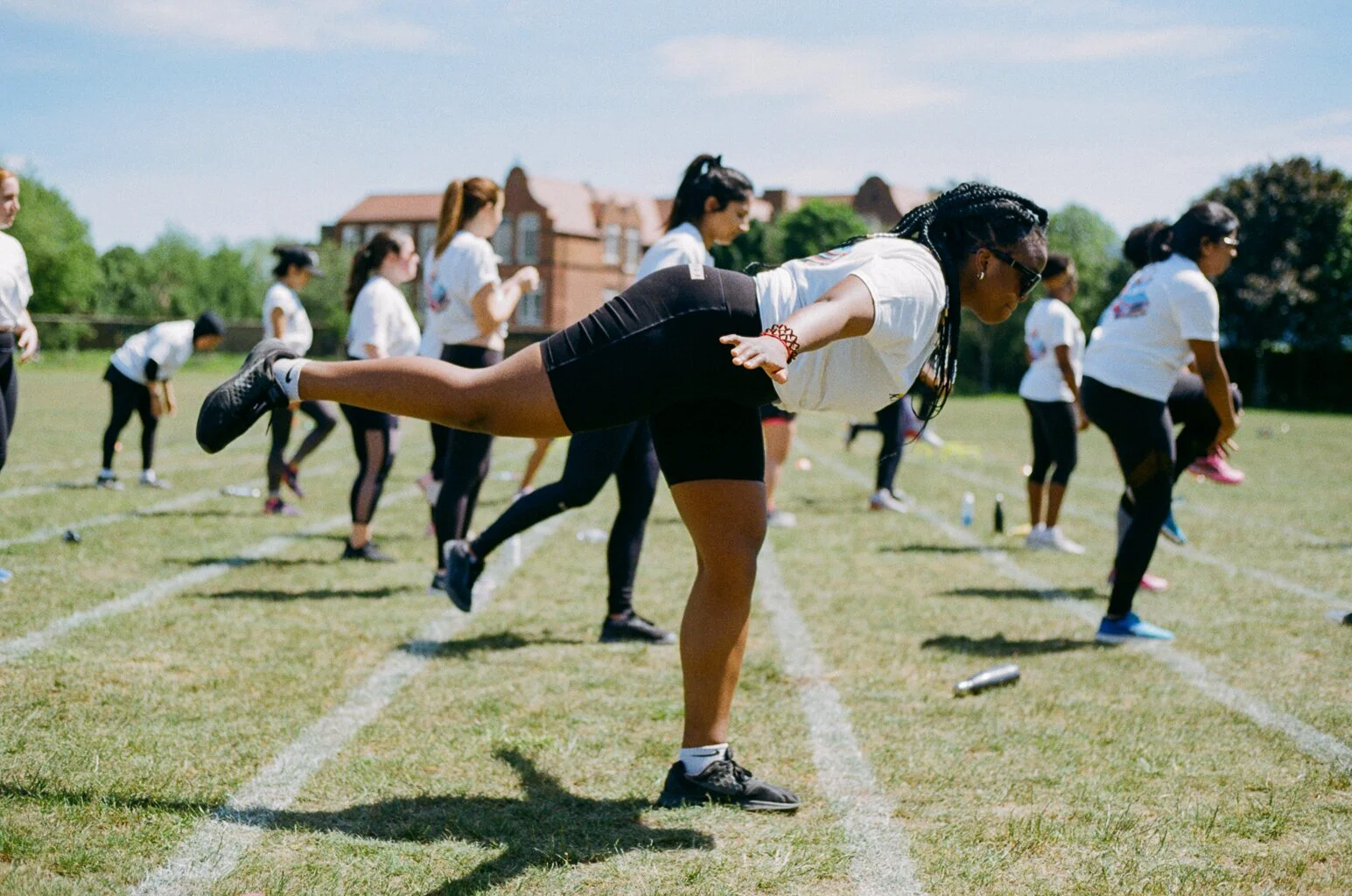 Group of women participating in an outdoor fitness class, with one woman in the foreground balancing on one leg with arms extended backward, on a grassy field on a sunny day.