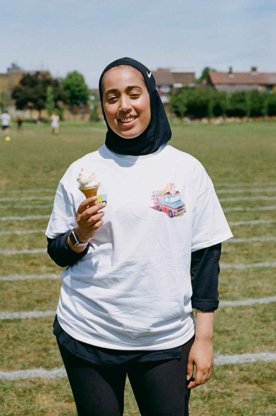 A woman wearing a black hijab and a white t-shirt holding an ice cream cone, standing on a grassy field with other people and trees in the background.
