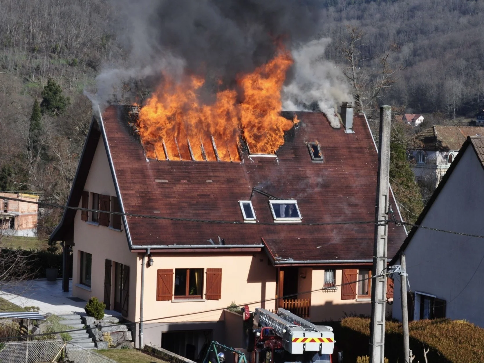 Un bâtiment résidentiel en flammes avec un toit en tuiles rouges, fumée et flammes s'échappant du toit, avec une échelle de pompiers en bas et des maisons environnantes en arrière-plan.