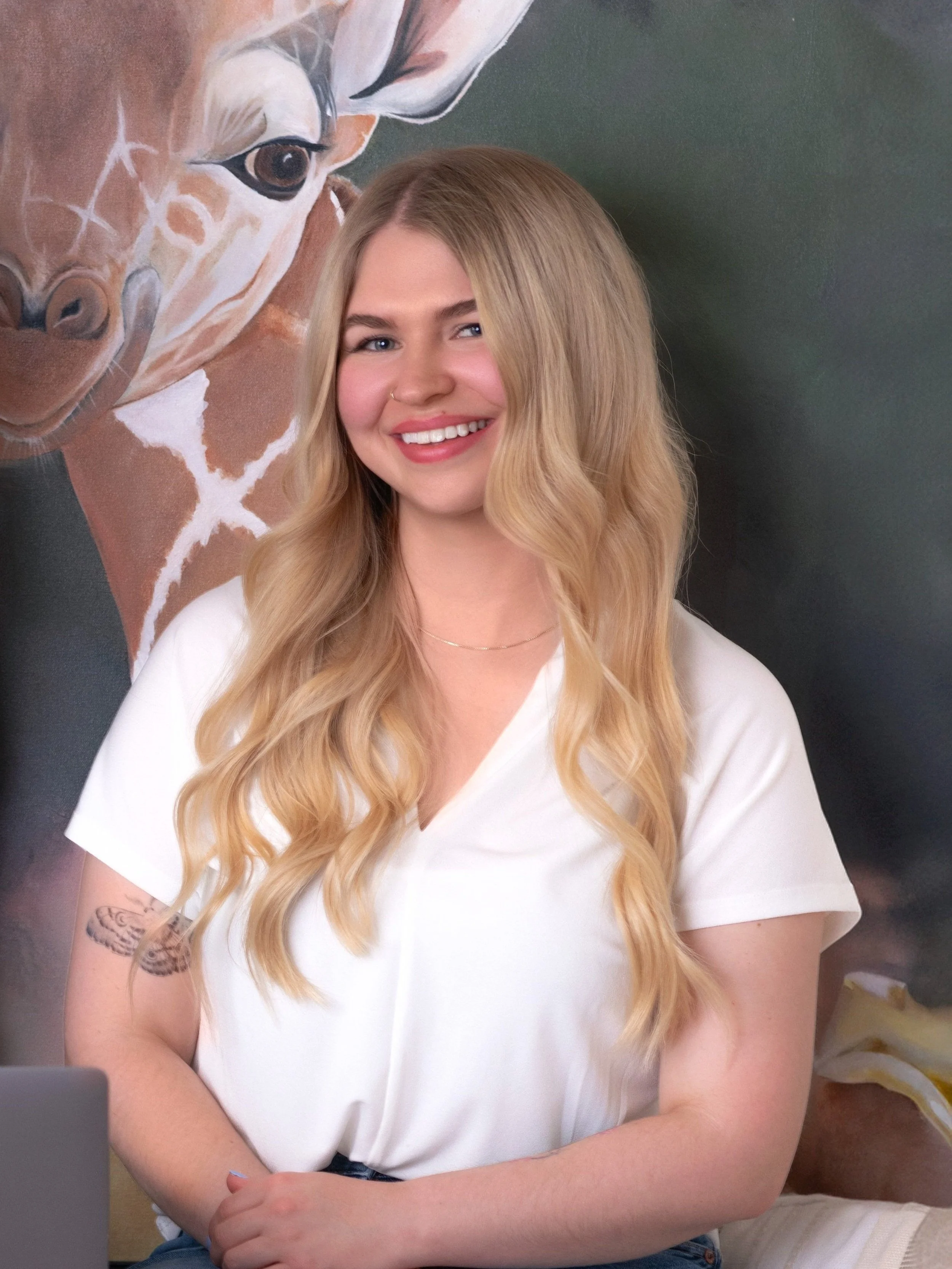Smiling woman with long blonde hair sitting in front of a giraffe painting, wearing a white shirt.
