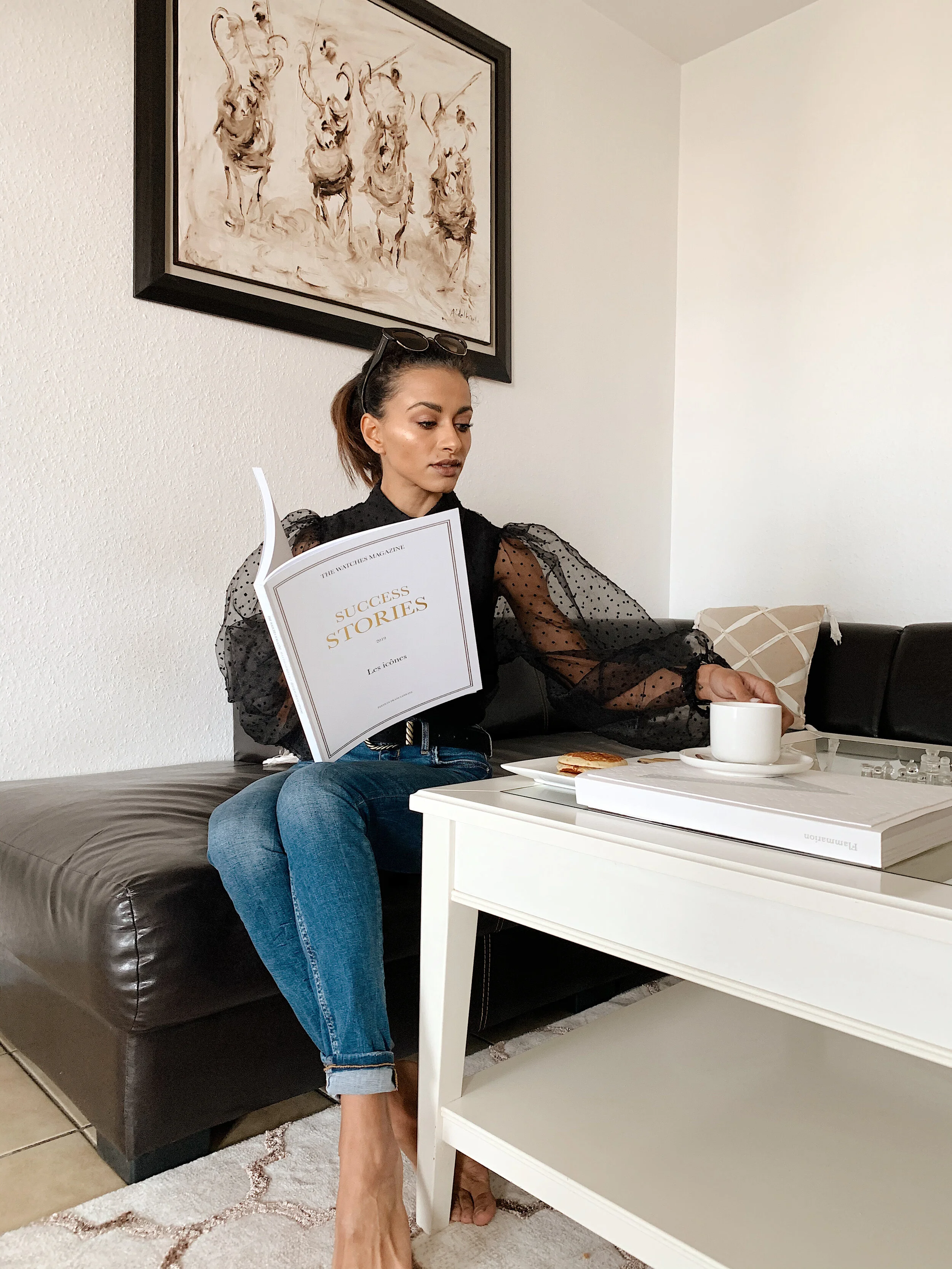 Diane GLAOUA sitting on a leather deep brown sofa, reading a magazine titled 'Success Stories' while reaching for a white coffee cup on a white table.
