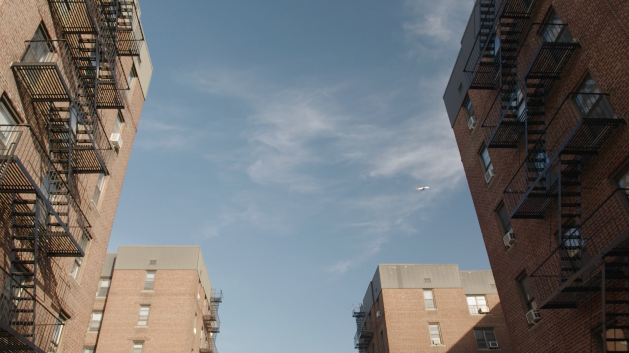 View of the sky between two red brick apartment buildings with fire escapes, and a small airplane flying in the distance.