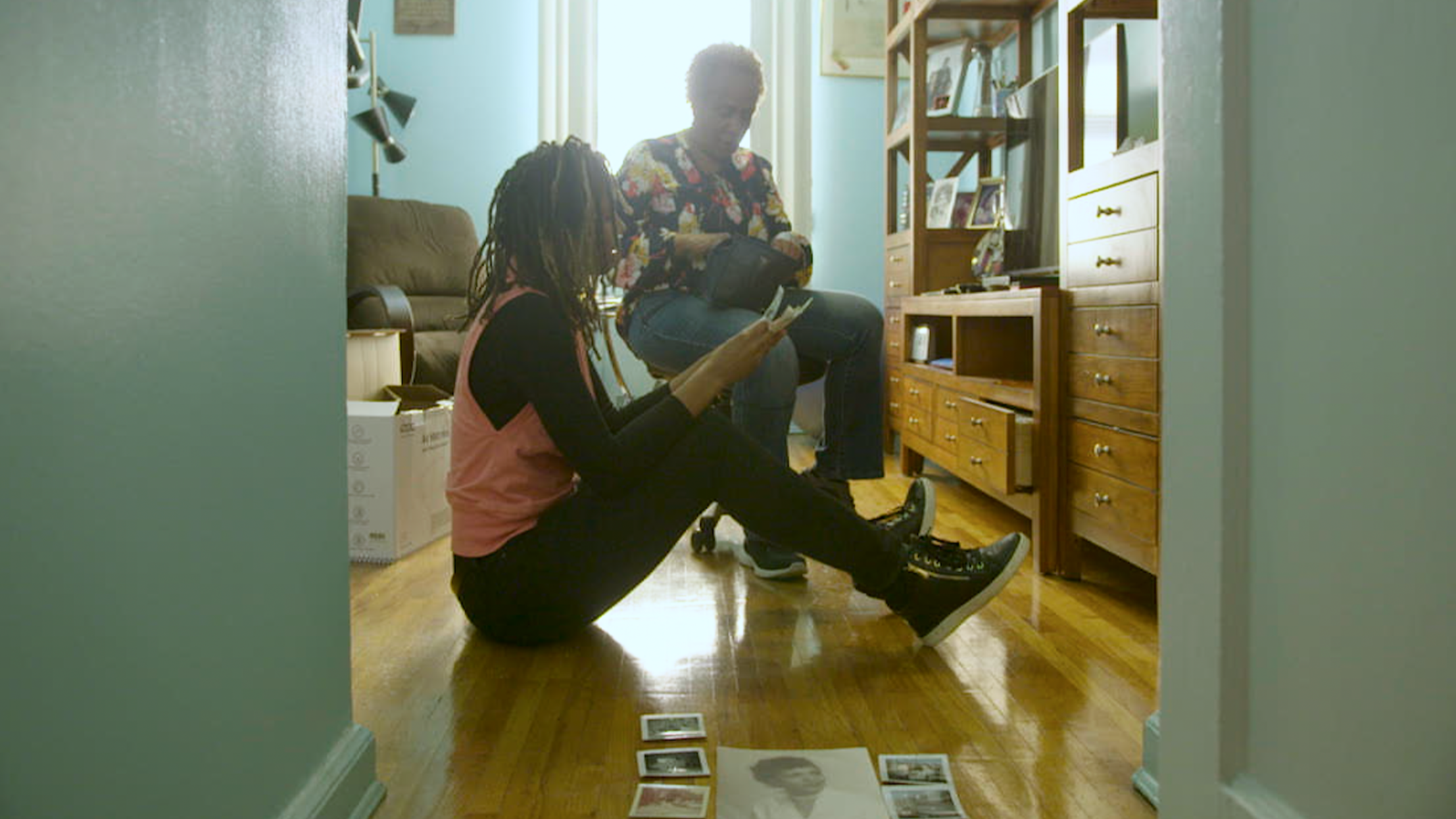 Two women with dreadlocks and curly hair in a living room, one sitting on the floor looking at photographs scattered on the wooden floor, and the other sitting on a couch, holding a photo, engaged in conversation. Sunlight streams in from a window be