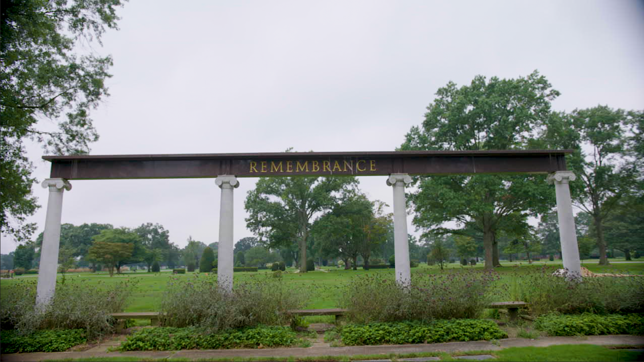 A park with trees and grass, featuring a large sign with the word 'REMEMBRANCE' on a wooden beam supported by white columns.