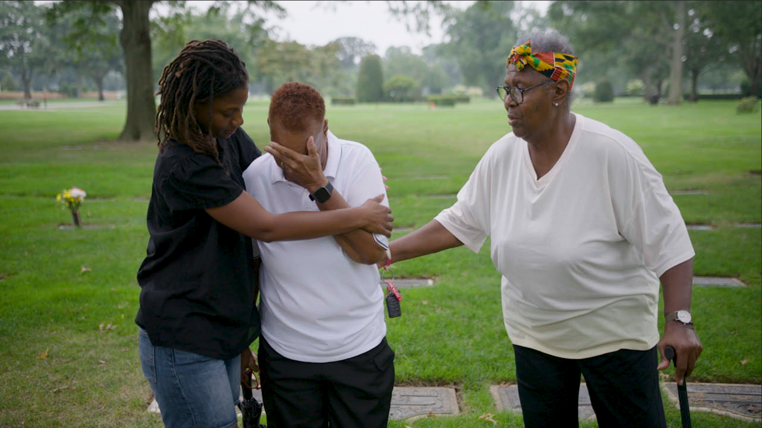 Three women standing outdoors in a park with trees, one appears distressed and is being comforted by the other two women.