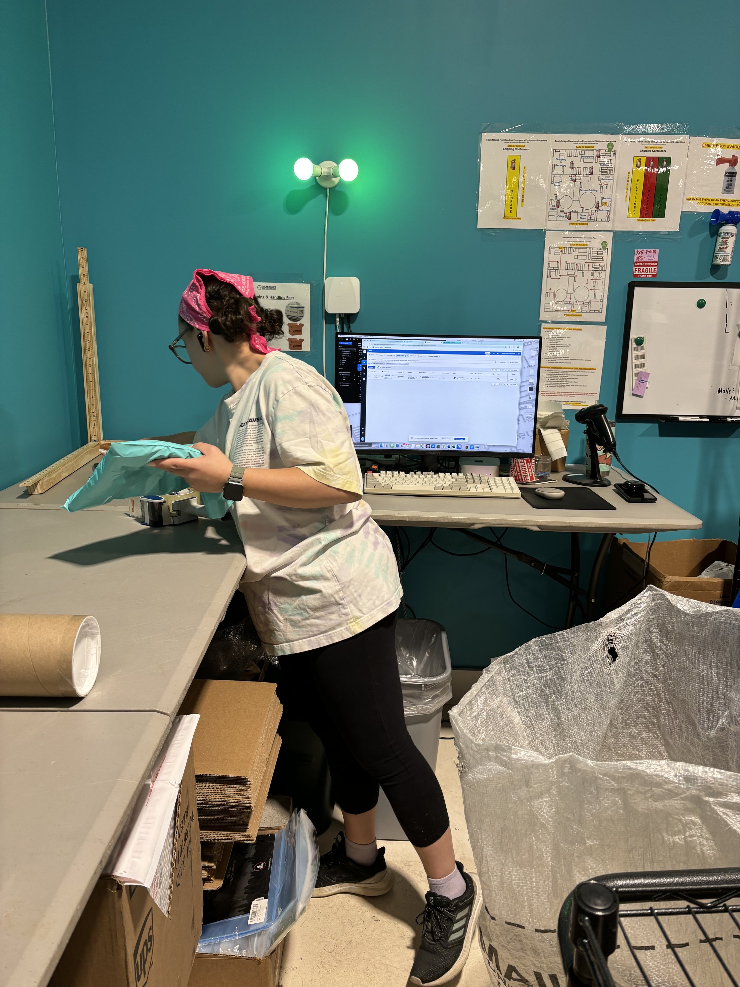 A woman wearing glasses, a pink headscarf, and a lab coat working at a cluttered workspace with a computer, monitor, and packaging materials in a teal-colored room with safety notices on the wall.
