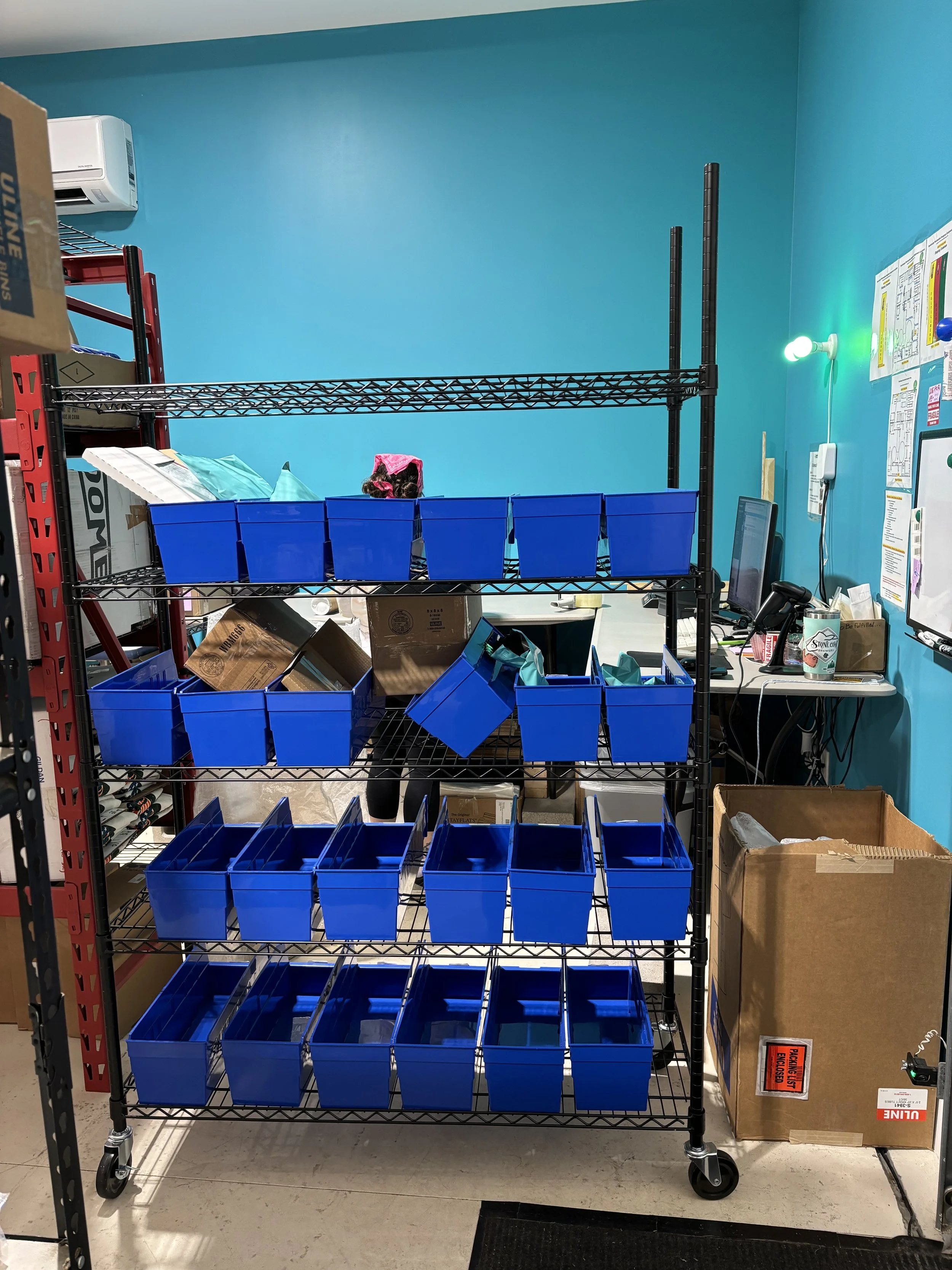 Metal storage rack with multiple blue plastic bins filled with supplies, in a workspace with turquoise walls, a computer, and office supplies on a desk.