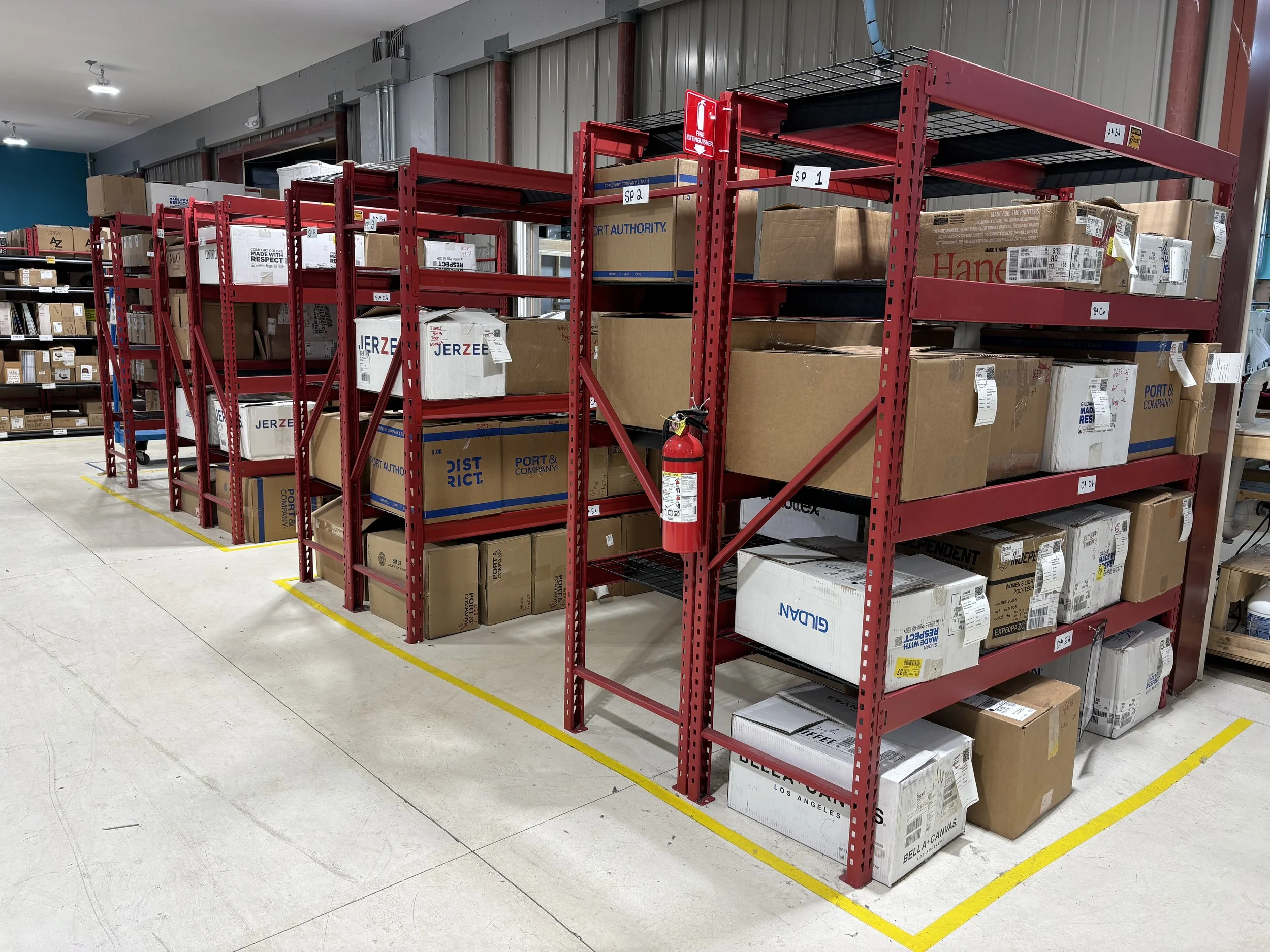 Warehouse storage with multiple red metal shelves filled with cardboard boxes and packages, fire extinguisher on the side of a shelf, and yellow safety lines on the floor marking the area.