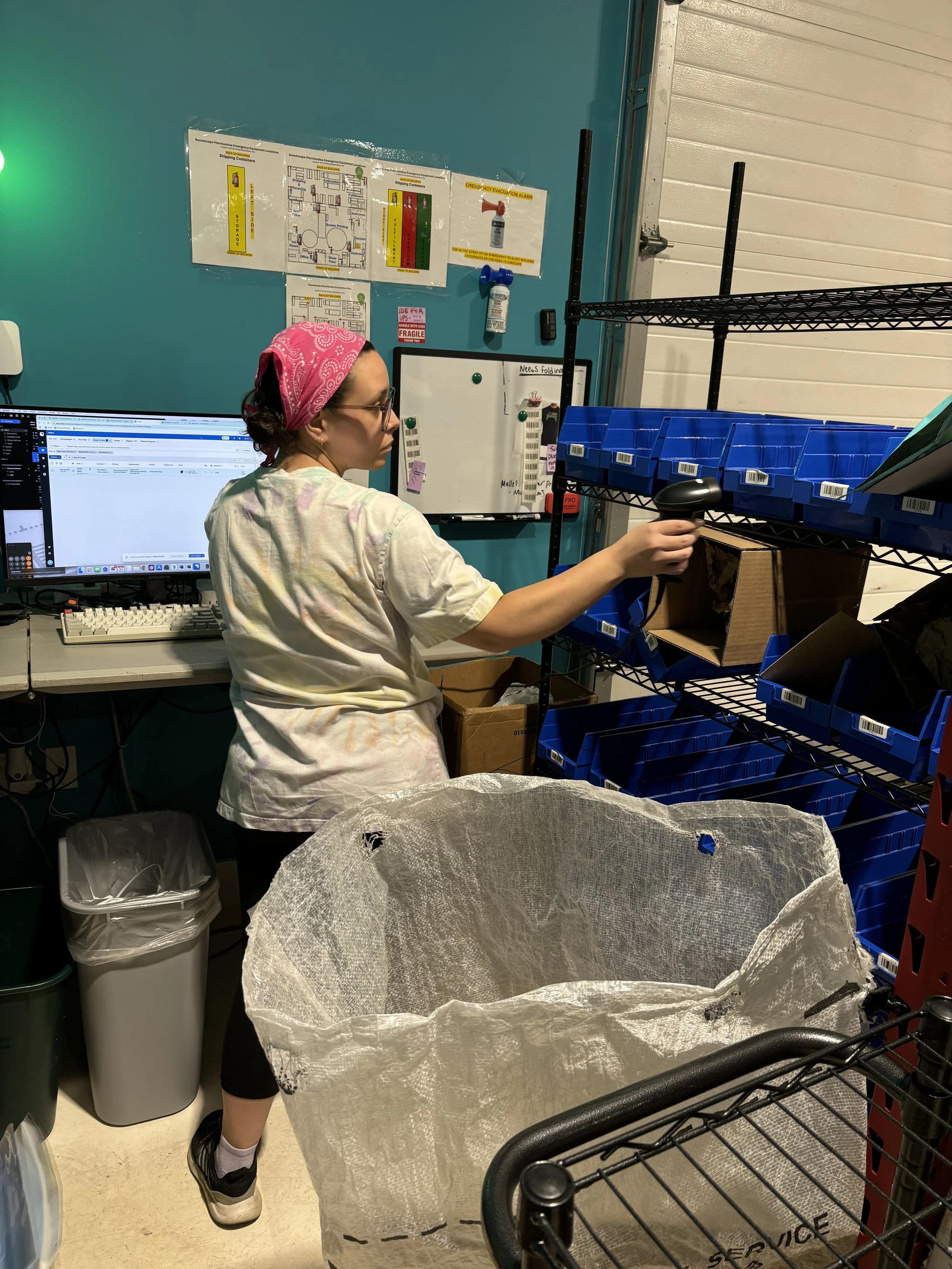 A woman wearing a pink bandana and glasses scans items on a metal wire shelving unit in a storage room with blue walls, a computer, and recycling bins.