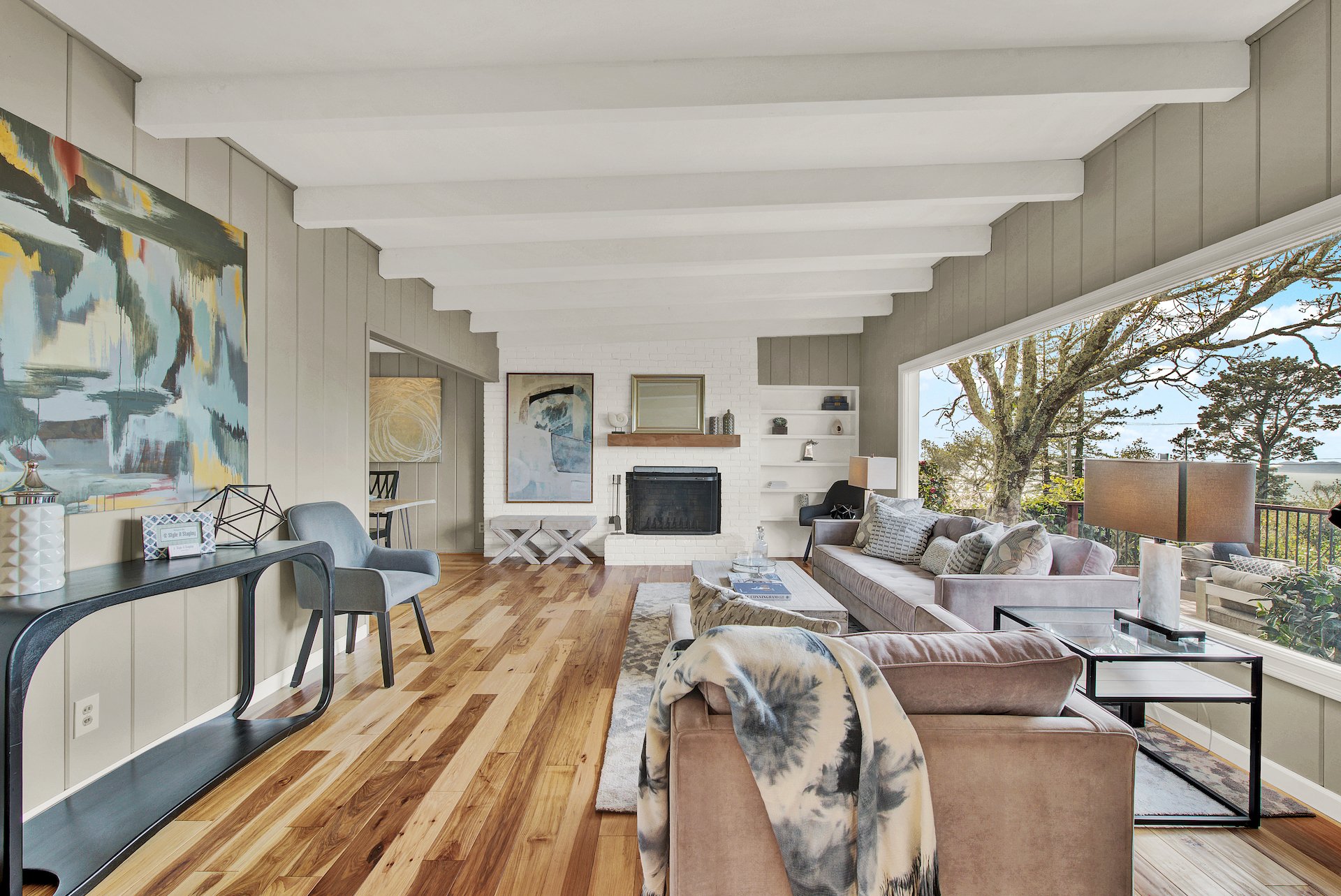 Living room styled for real estate sale. Large picture window, freshly painted camel colored walls, Brick fire place painted white, Ceiling wood beams painted white. Modern art and furniture. Designer styled by Style it Staging