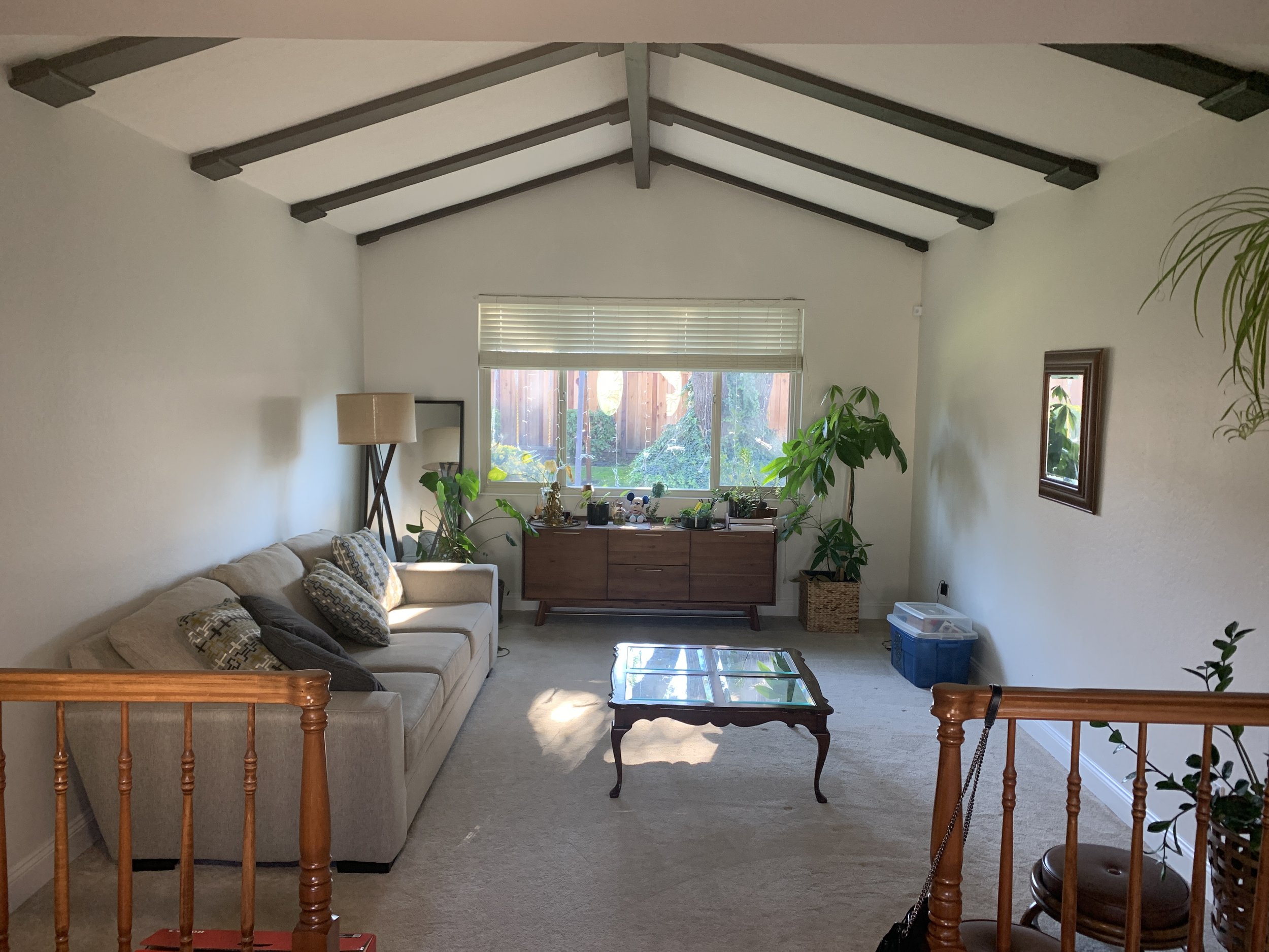 Before styling step down living room with wood banister and vaulted white ceilings with dark wood beams. Furniture is bulky and oversized