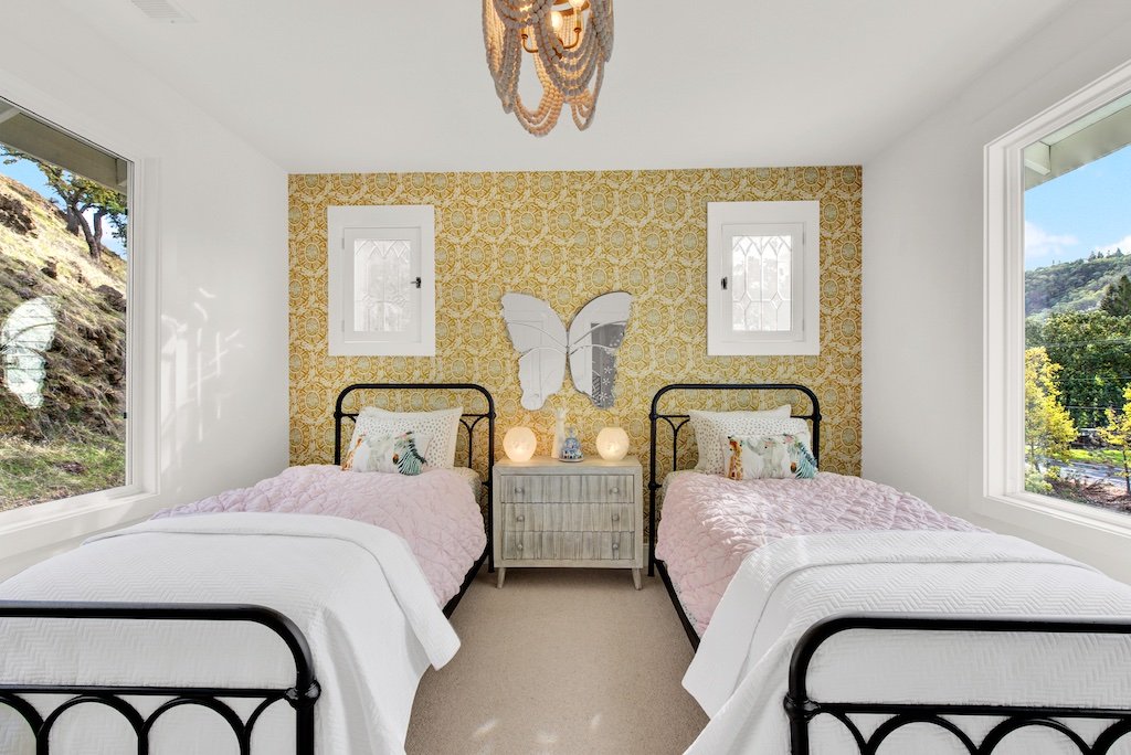 Little girls bedroom with ornate gold wallpaper on back wall and beaded chandelier. 2 twin beds with wrought iron bed frames.