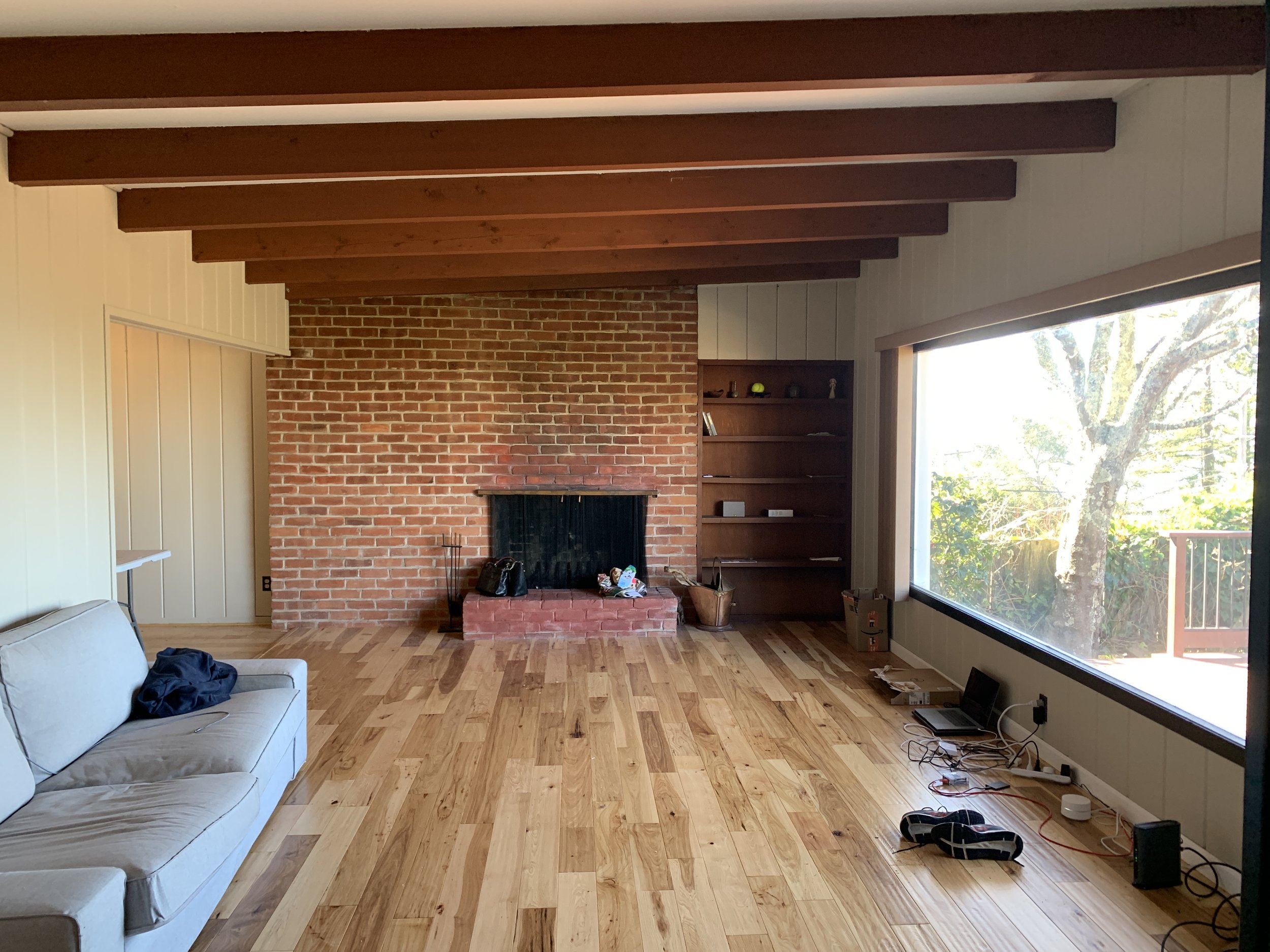 Living room with large window, wood floors, brick fire place, wood beams on ceiling. One broken couch in an otherwise empty room.
