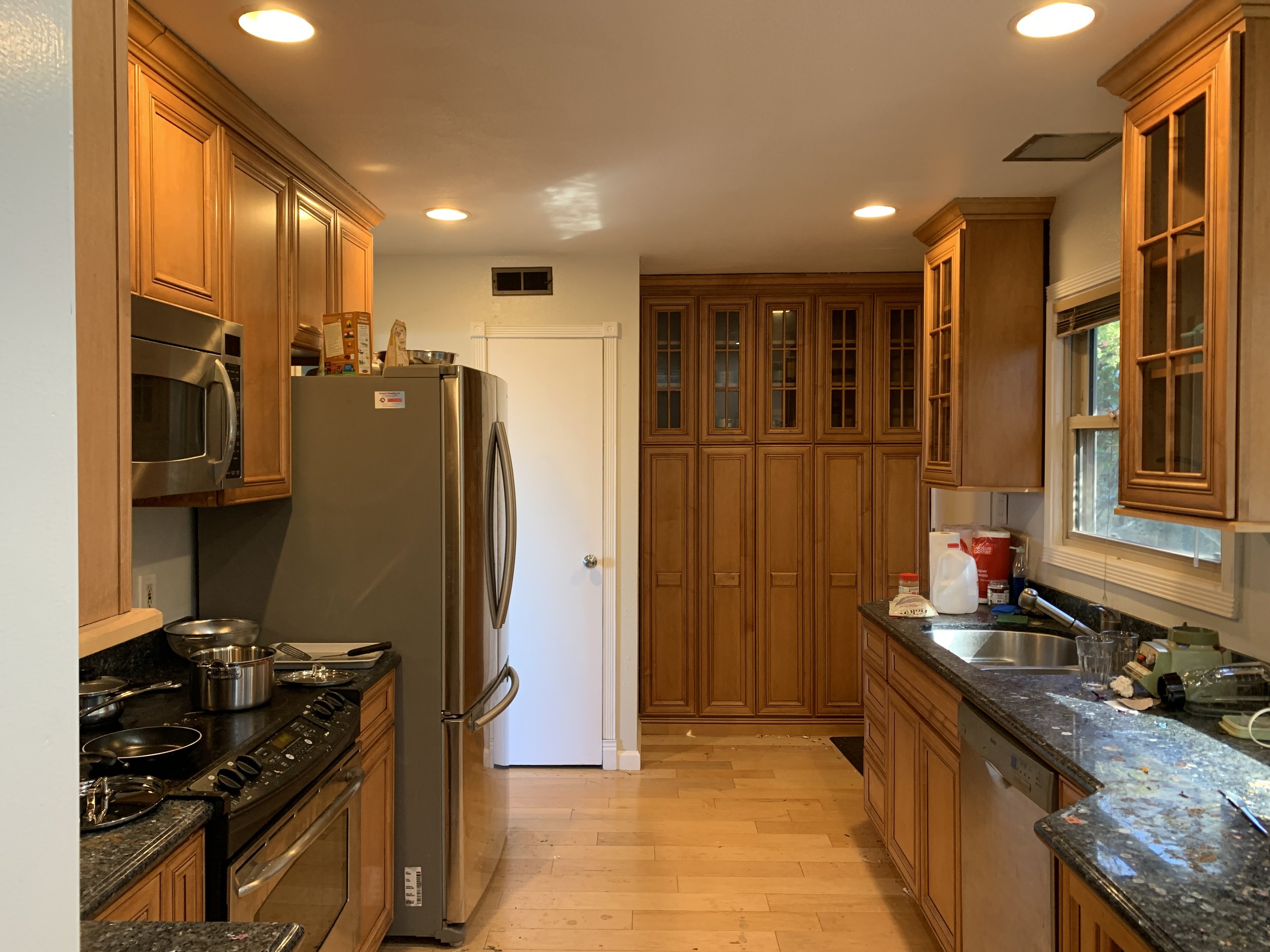 Galley style kitchen with medium stained wood cabinets and black granite counter tips. Wood pantry on back wall. Before photo in a before and after series