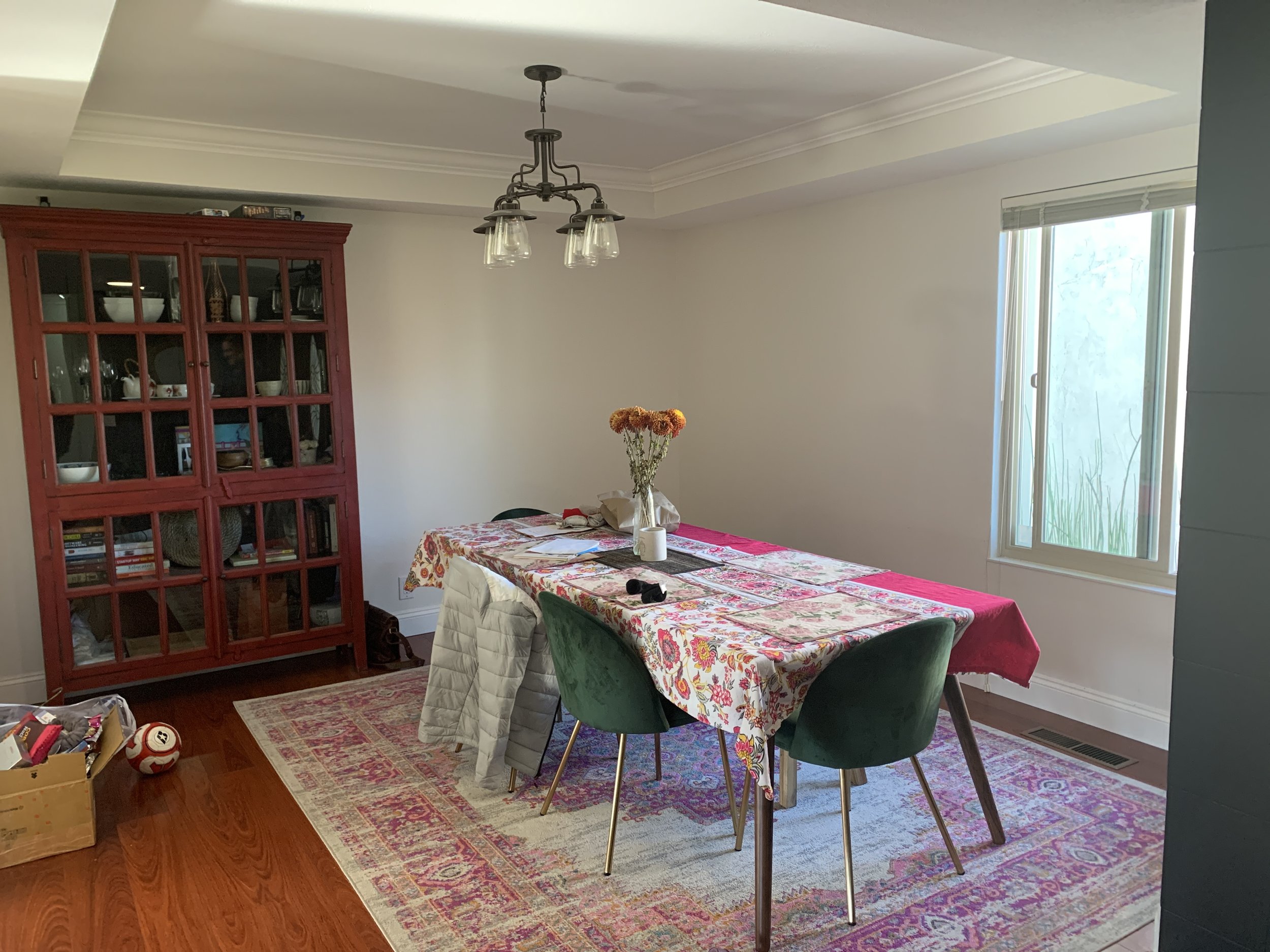 Home staging before photo of a dining room. Floral table cloth and large hutch in corner and area rug underneath the table. Makes the room look a little dumpy. Beautiful coffered ceiling and large window.