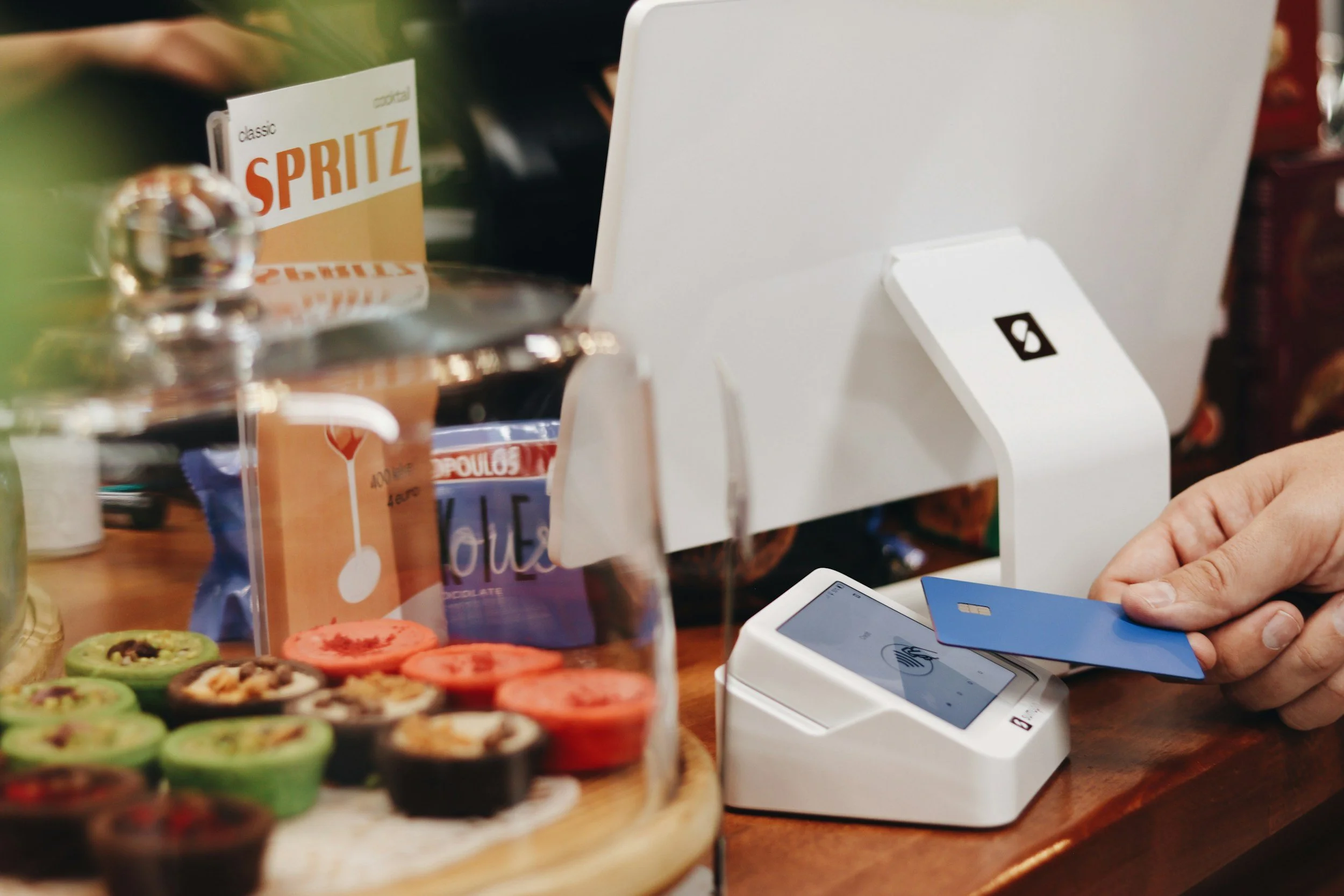 Person holding a blue credit card at a white point-of-sale terminal, with a large white display screen, on a wooden counter. In the foreground, there are various colorful cookies on a tray, and behind it, snack packages including a Sprite can and a chocolate bar.