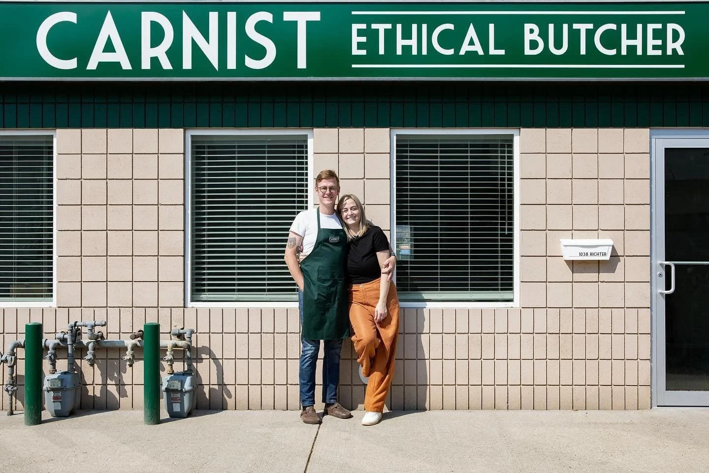 Two people smiling outside an ethical butcher shop called CARNIST, one wearing an apron, standing close together in front of the shop's window.