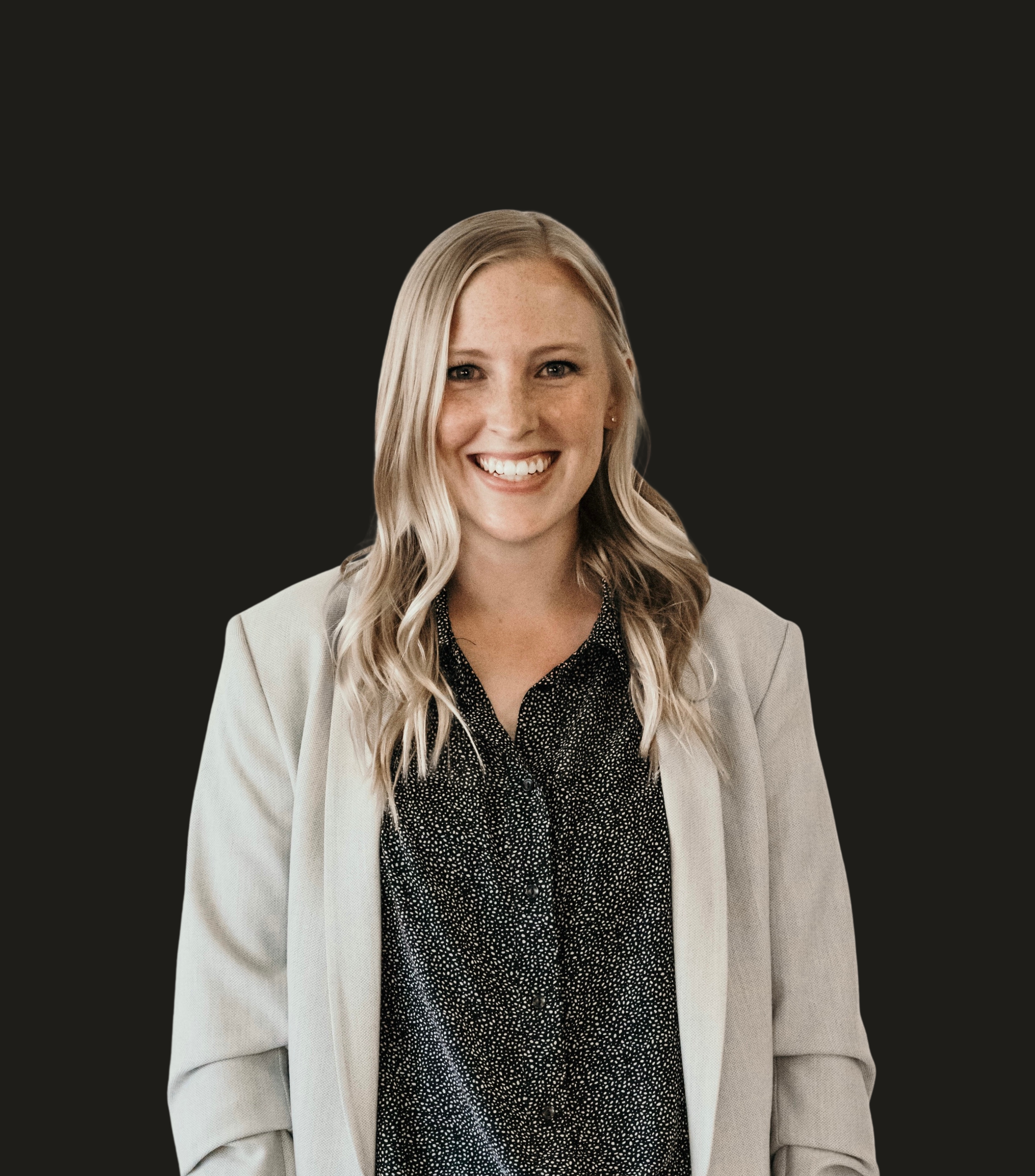 A smiling woman with long blonde hair wearing a beige blazer and a black blouse with white specks, against a dark background.