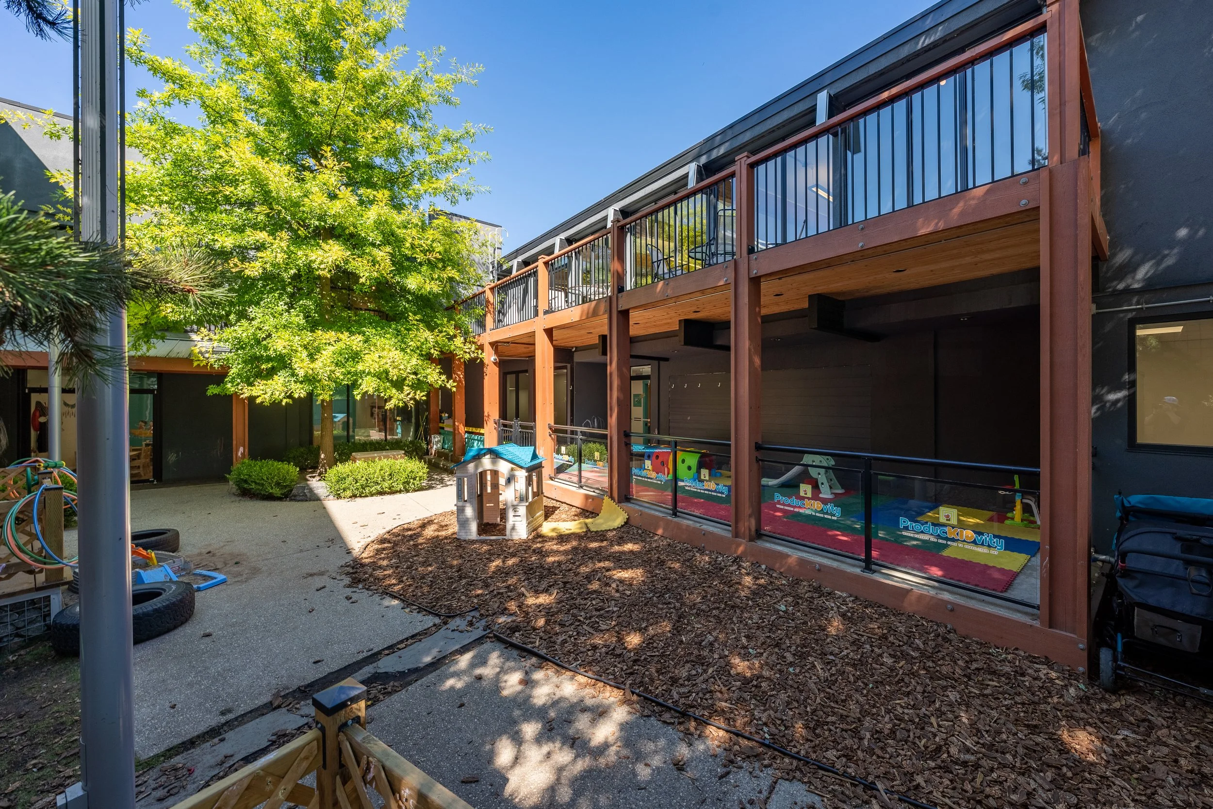 Outdoor playground area with a small playhouse and play equipment, surrounded by greenery and a modern building. Sunny day with clear skies.