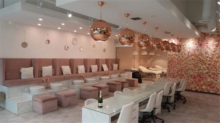 Spa lounge area with pink seating, white towels, and rose gold pendant lights, including pedicure stations and floral wall decor.