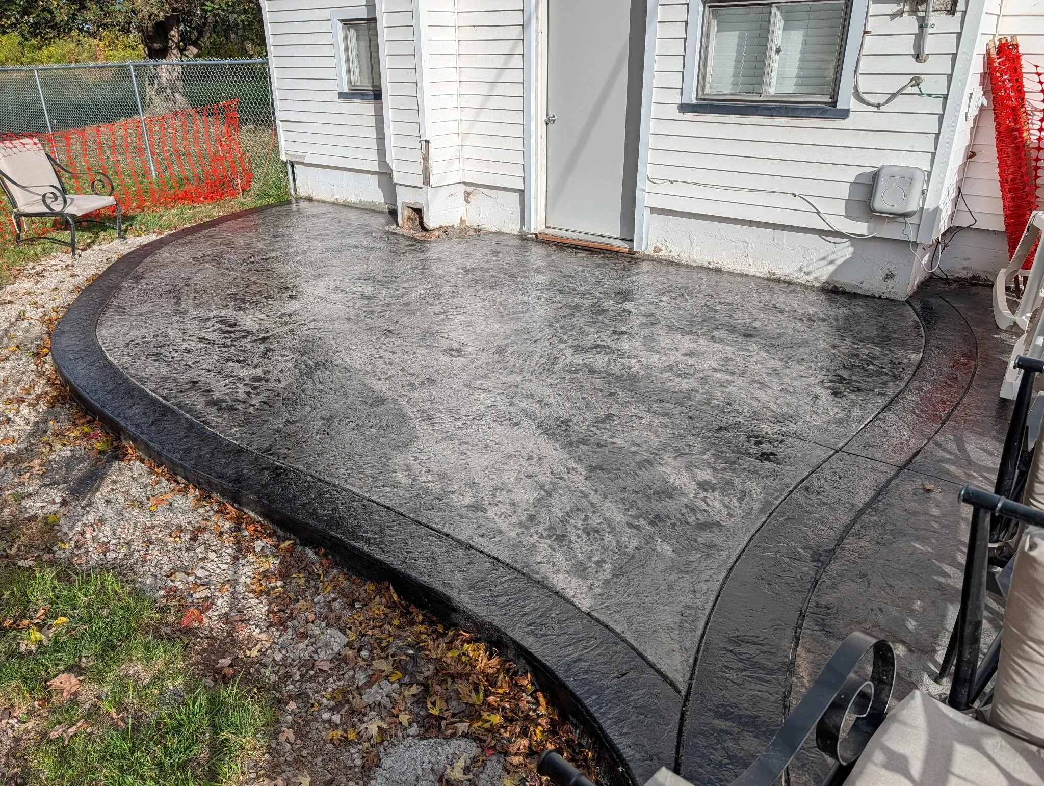 Freshly poured concrete patio outside house with white siding, a closed door, window, and nearby outdoor furniture, including a bench and red plastic fencing.