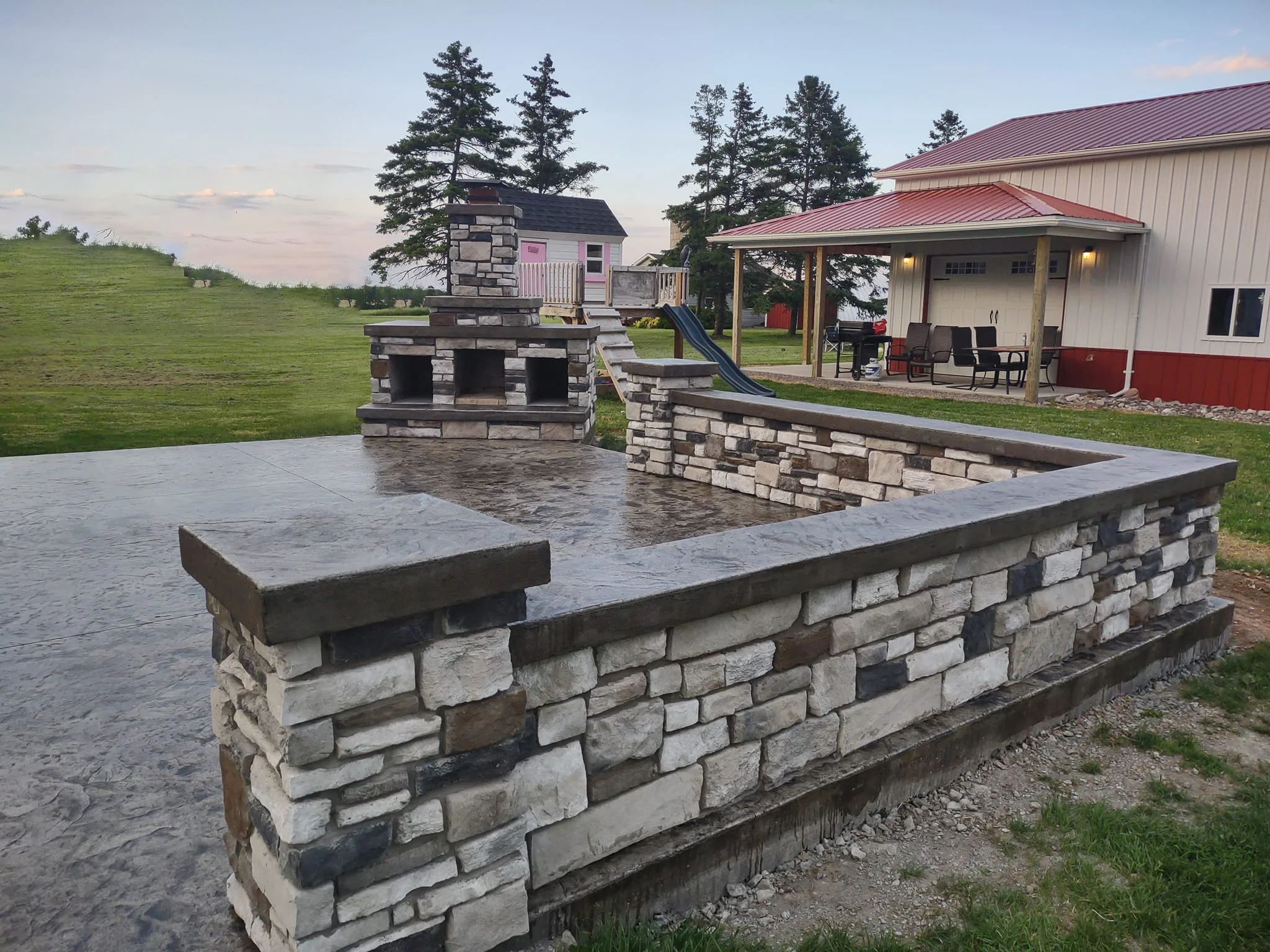 Outdoor stone patio with built-in grill and chimney, surrounded by a low stone wall, backyard with grass, trees, a house with a porch, and outdoor furniture.