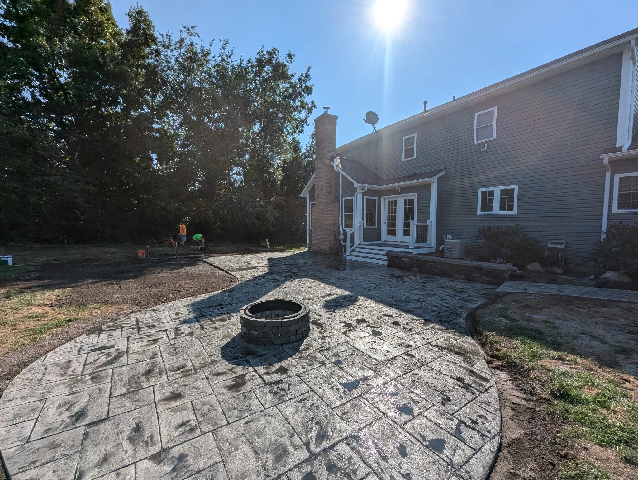 Backyard with newly laid patterned stone patio, a small firepit, and a house with a brick chimney and sliding glass doors; two workers in the distance are working on landscaping, with trees on the left and the sun shining brightly overhead.