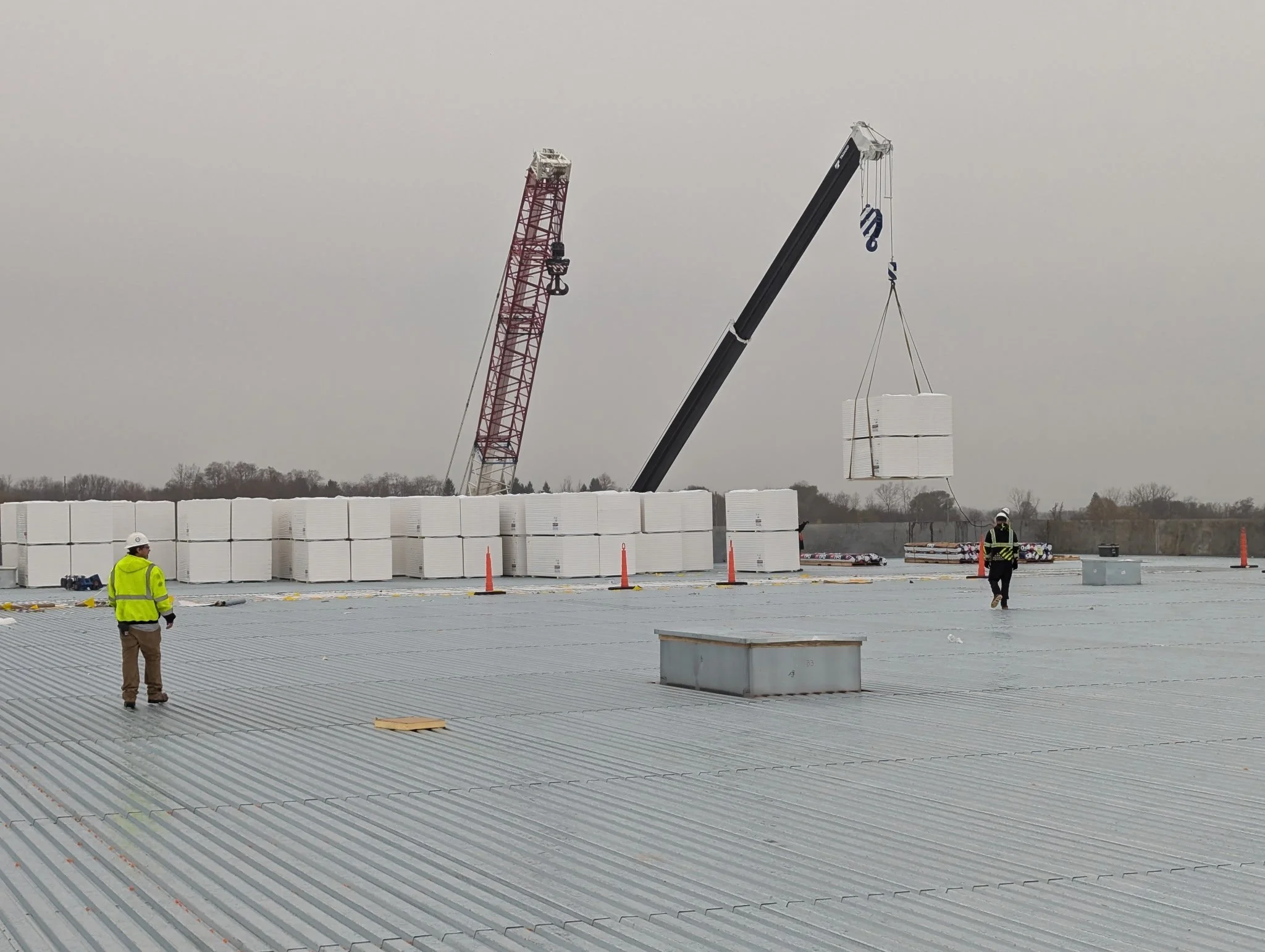 Construction site on a warehouse roof with two workers, one standing and one walking, and a crane lifting palletized materials.