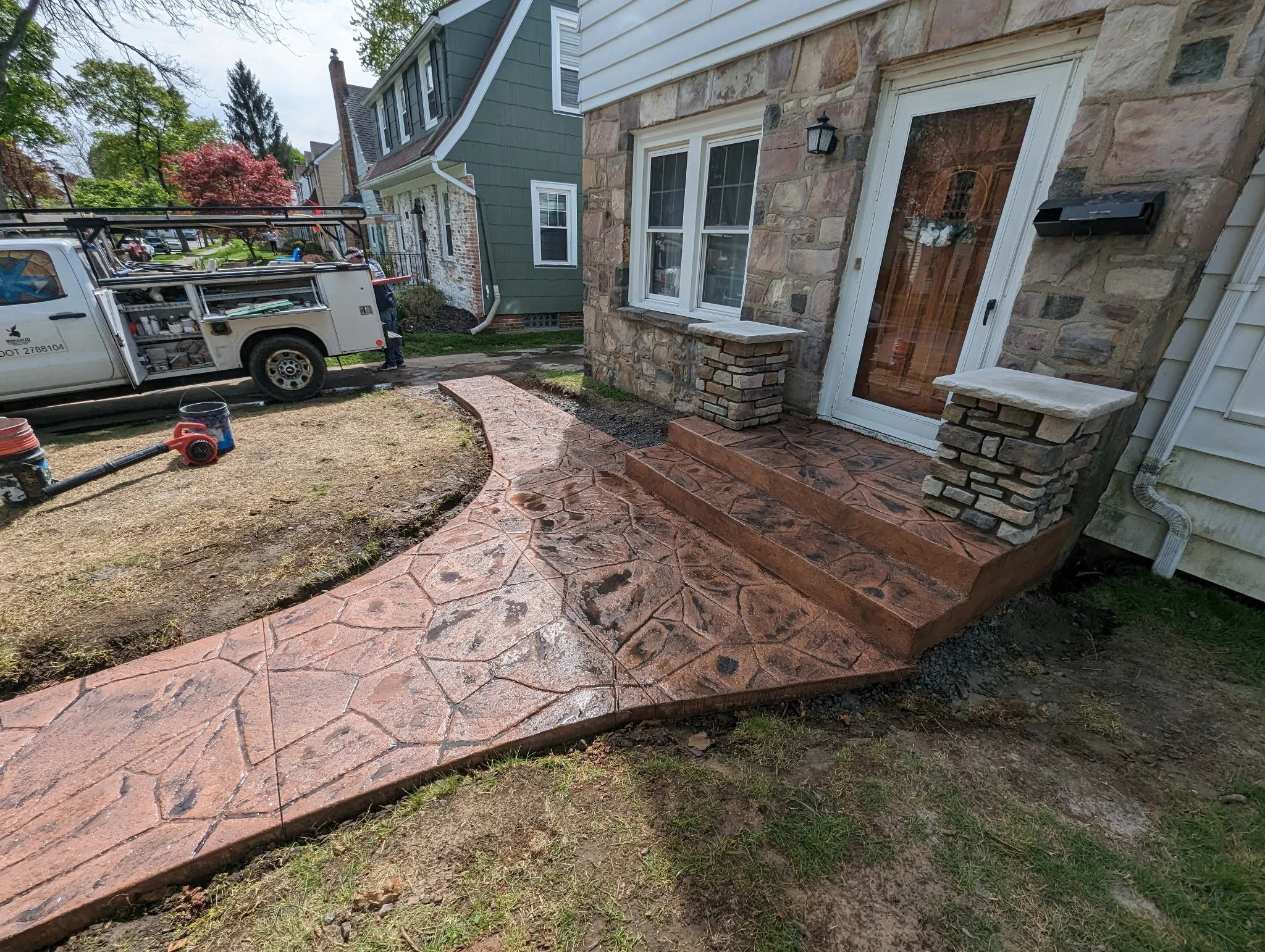 Front porch with a decorative stone walkway and steps leading to a glass door. A black mailbox is mounted near the door and a stone accent wall with bricks is visible. A white utility truck is parked nearby, and the yard appears to be under landscapi