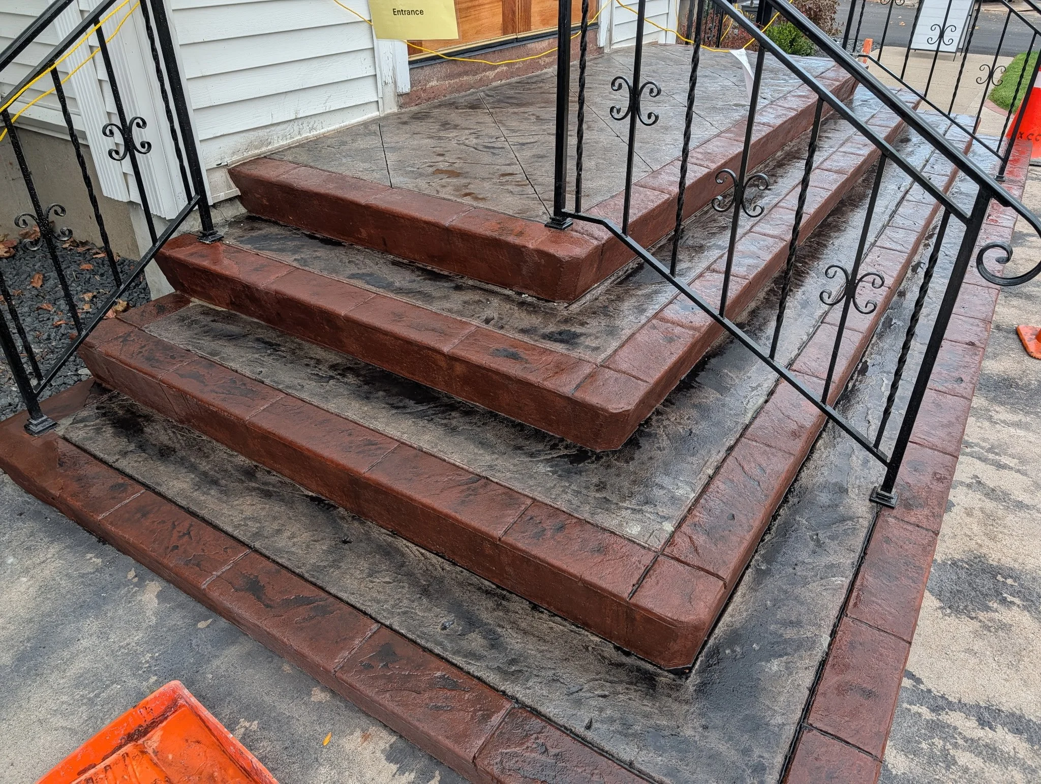 A set of brick stairs with three steps leading up to a porch area. The stairs are newly constructed and appear to be wet, with some water runoff. The porch has a black metal railing with decorative scrollwork. There is a yellow sign that says 'Entran