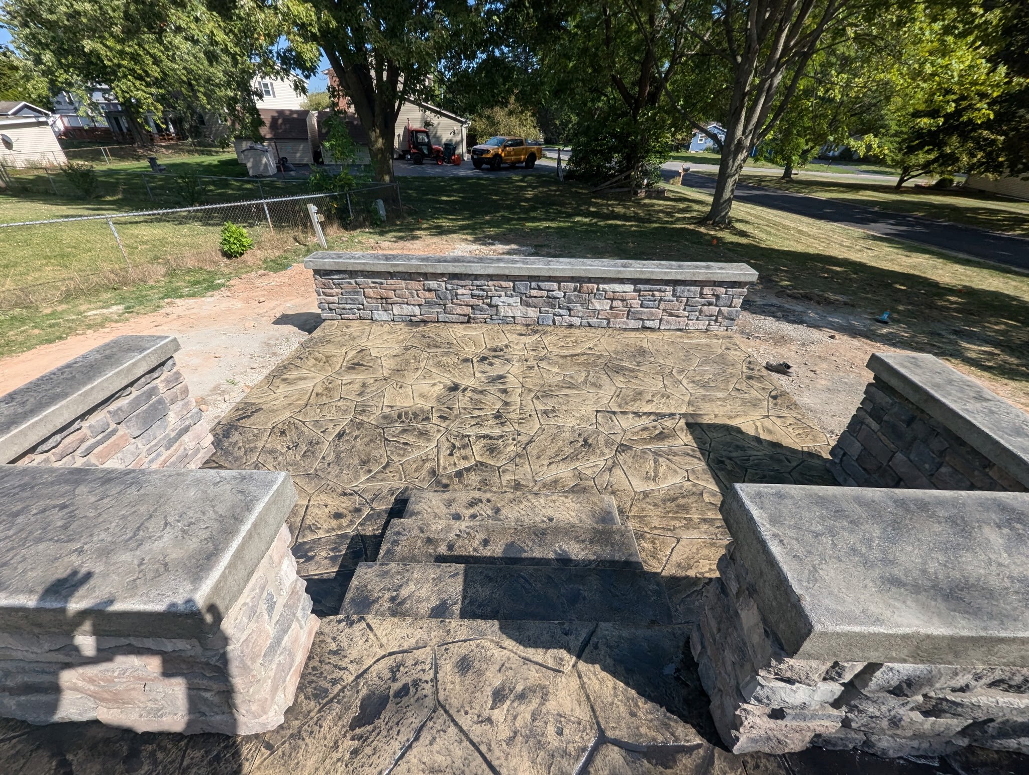 Front view of freshly constructed stone and concrete patio with steps, surrounded by a low stone and brick wall on a residential street with trees and houses in background.