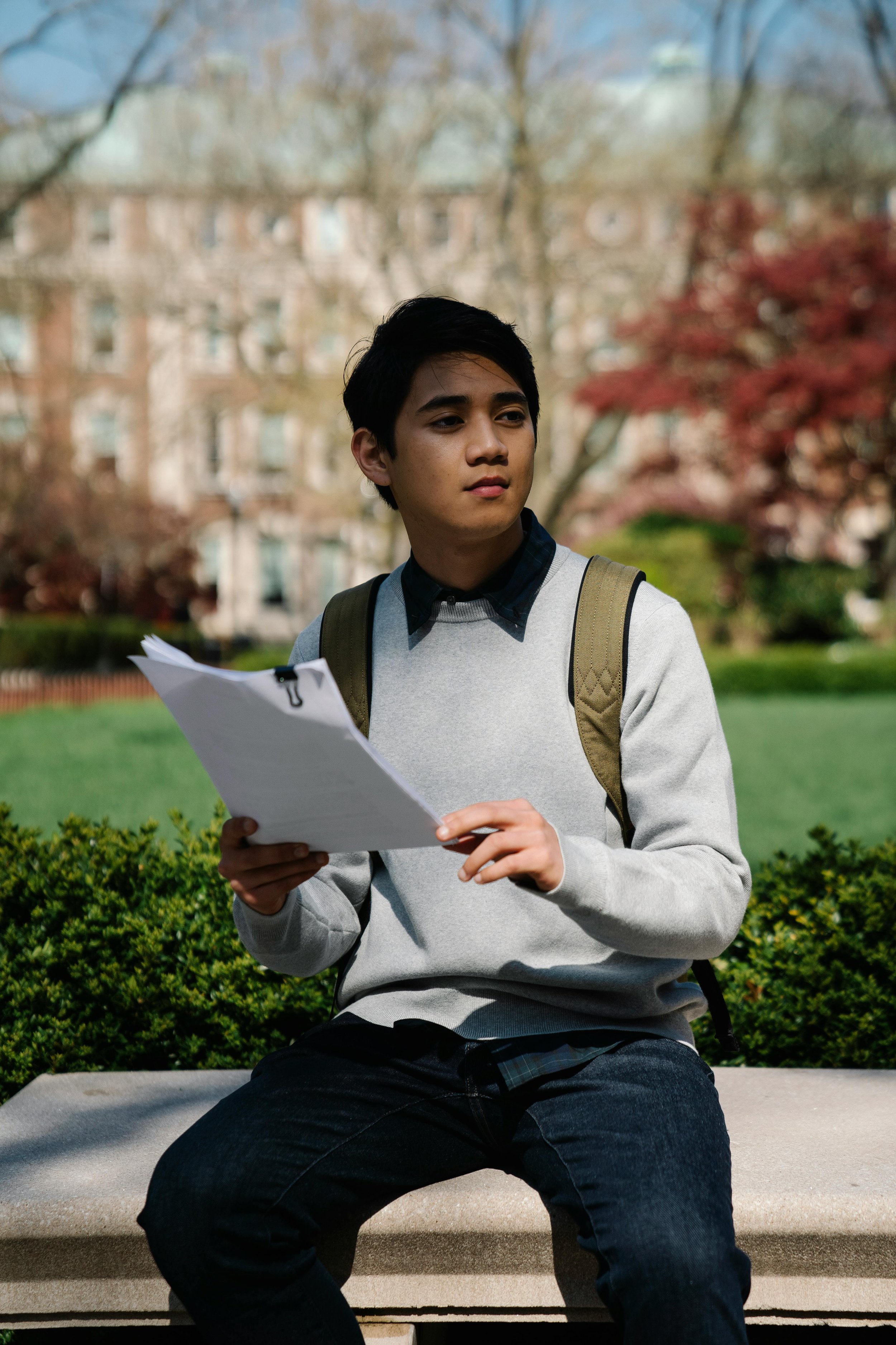 A college student sitting outdoors on campus, reviewing papers and looking thoughtfully ahead.