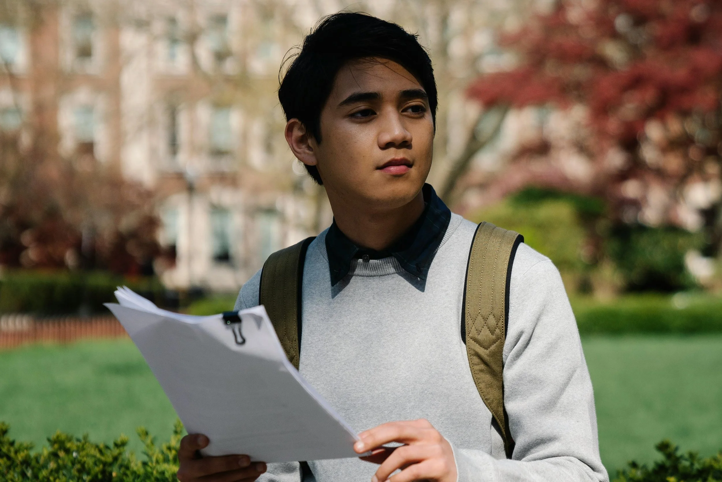 A college student sitting on a campus bench reviewing documents, looking thoughtful