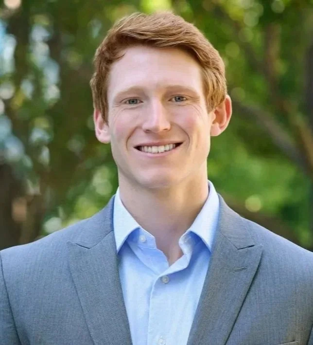 A young man with red hair and a bright smile, wearing a light blue shirt and gray suit, standing outdoors with a background of green trees.