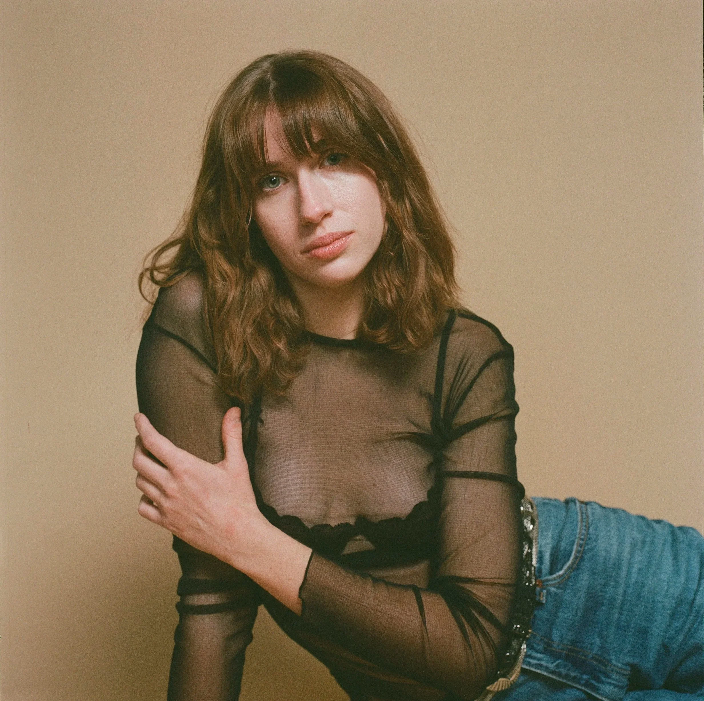 A woman with wavy brown hair and fair skin, wearing a sheer black long-sleeve top and blue jeans, posing against a plain beige background.