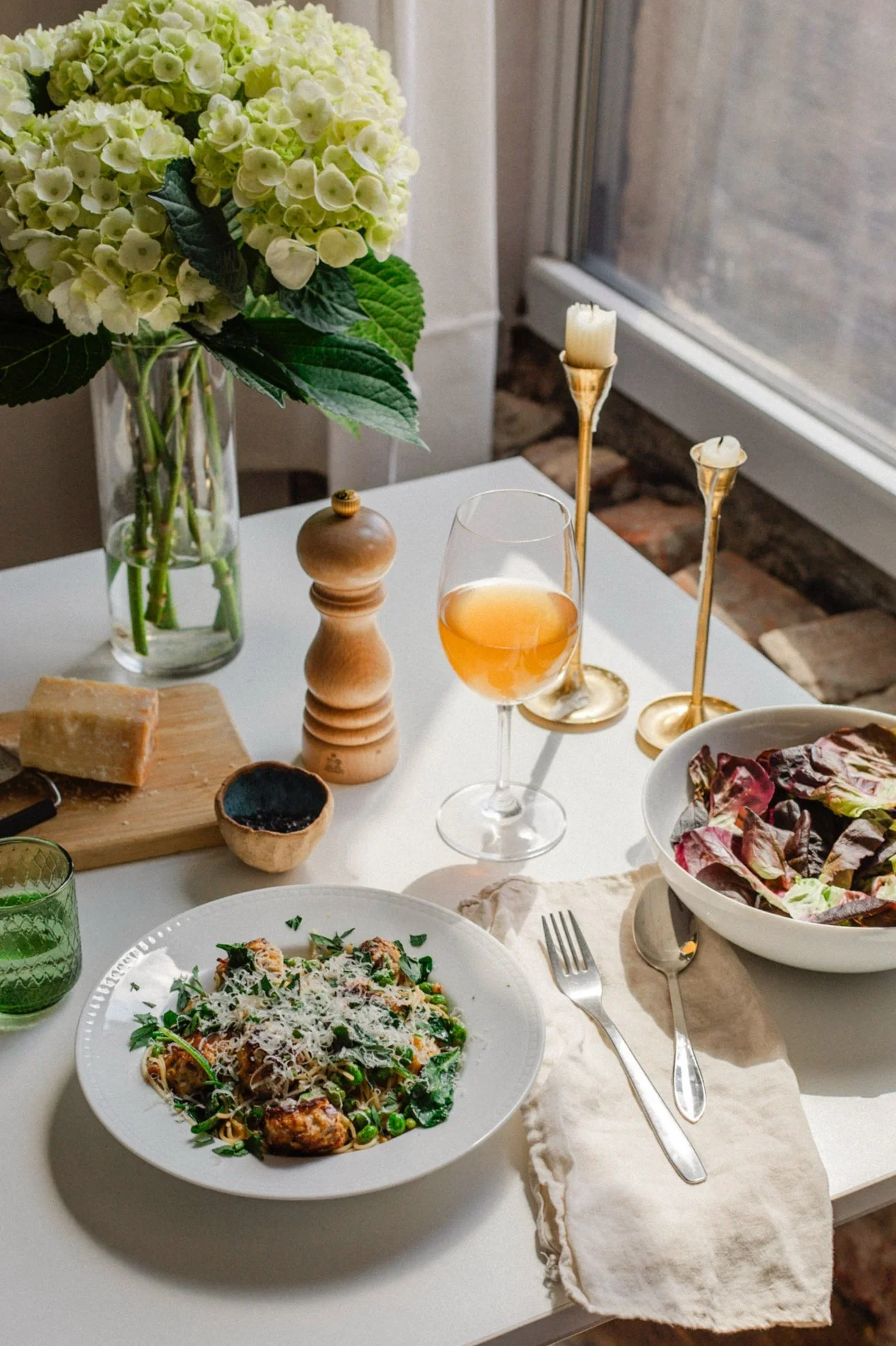 A white table set with a plate of spaghetti with greens and grated cheese, a side bowl of salad greens, a glass of rosé wine, lit candles, a vase of white hydrangeas, a wooden pepper grinder, a piece of cheese on a cutting board, and a small bowl of 