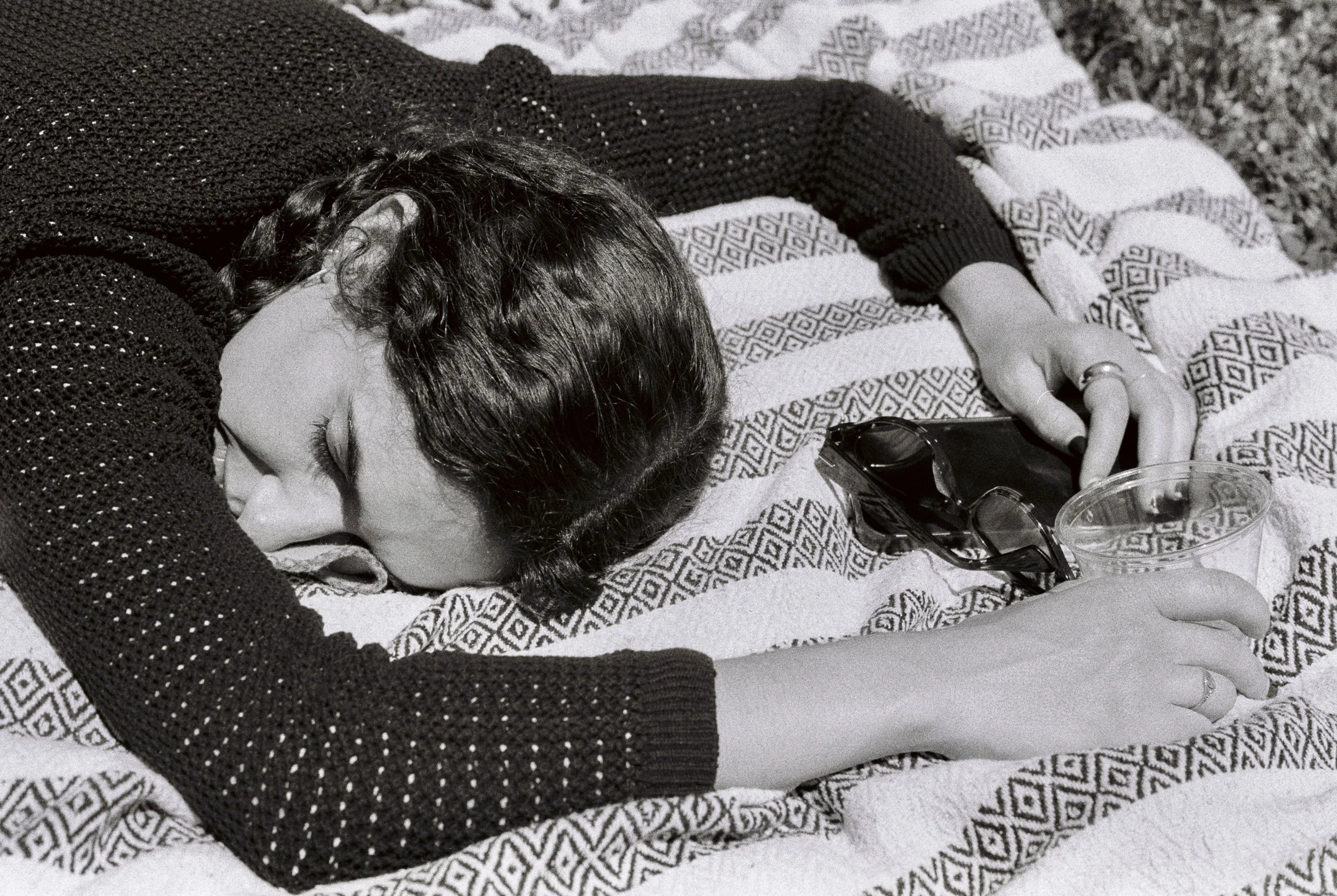 A woman with short dark hair lying face down on a patterned blanket, holding a plastic cup and a pair of sunglasses, appearing to be asleep.