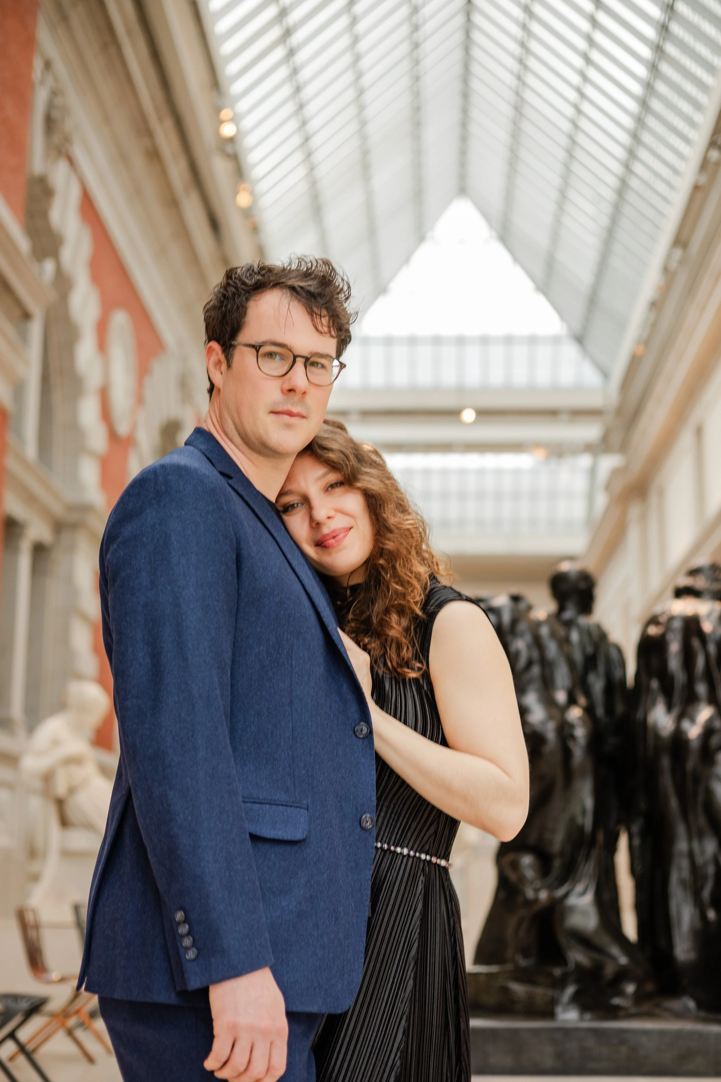 A man and woman hugging inside an art gallery or museum, with sculptures and statues in the background.