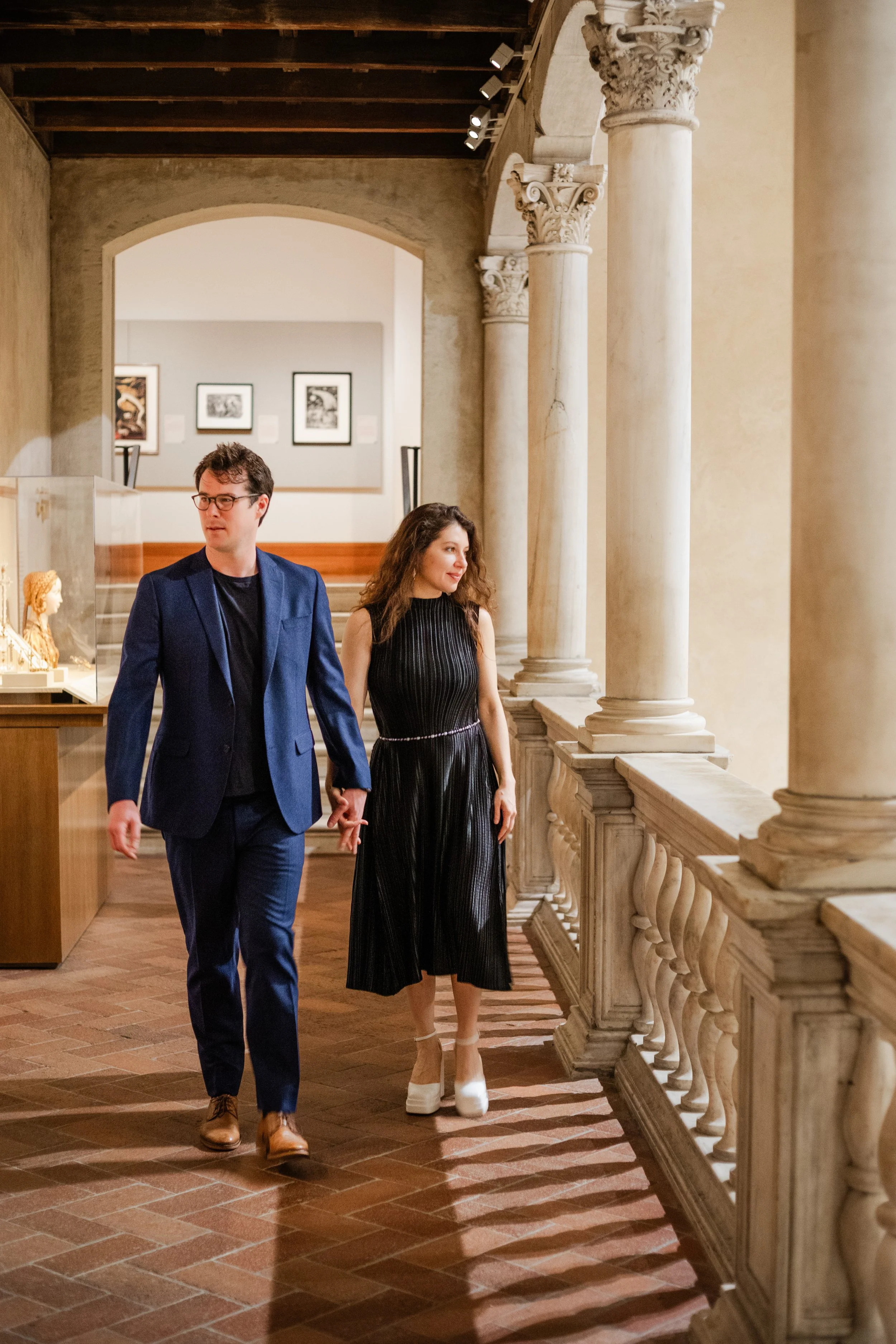 A man and a woman walking together inside a museum. The man is dressed in a blue suit with brown shoes, and the woman is wearing a black dress with white shoes. They are holding hands and looking at each other. The museum features classical architect