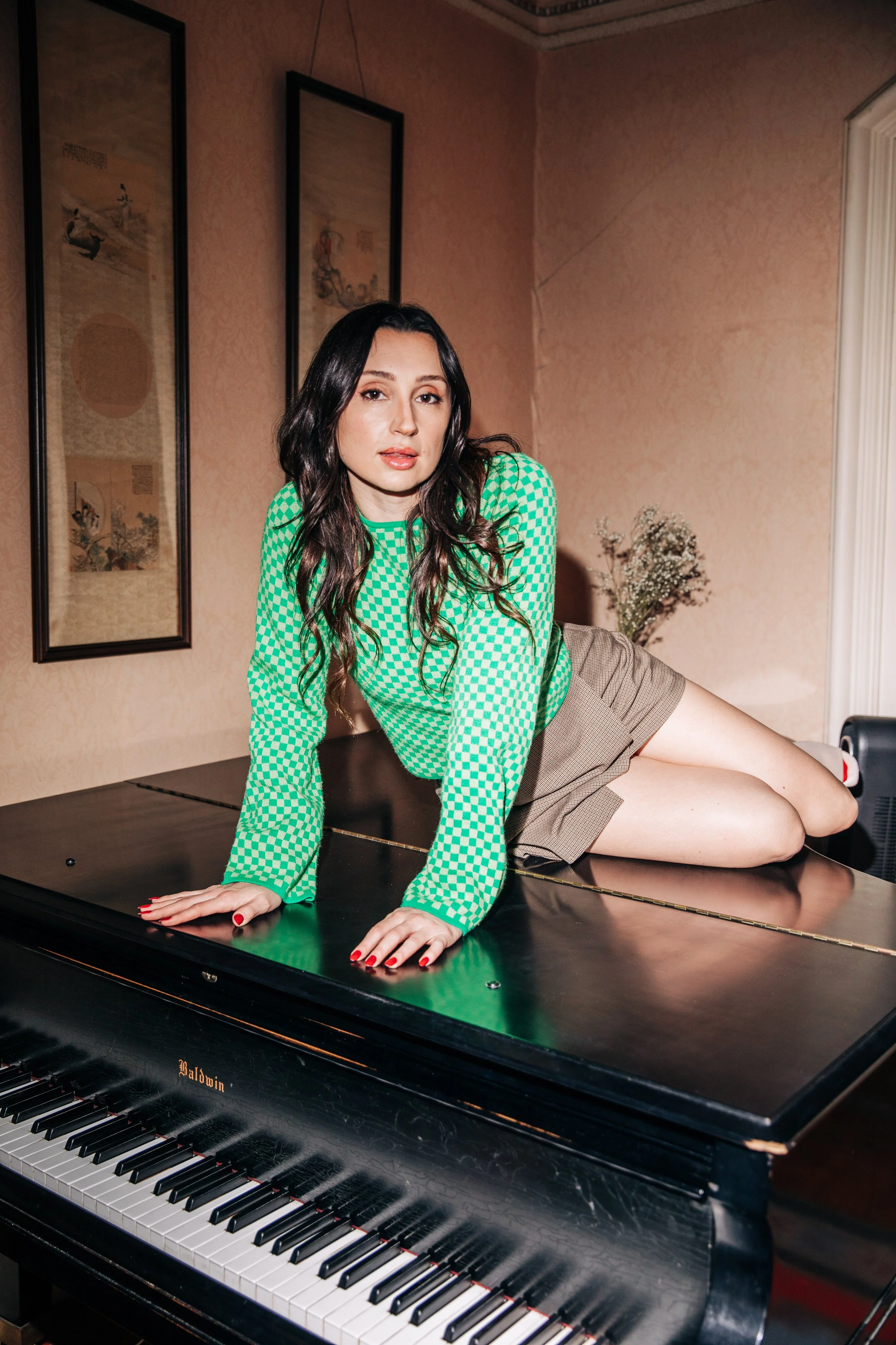 A woman with long wavy dark hair wearing a green checkered long-sleeve top and brown shorts, lying on top of a black Baldwin grand piano in a room with pink patterned wallpaper and framed artwork on the wall.