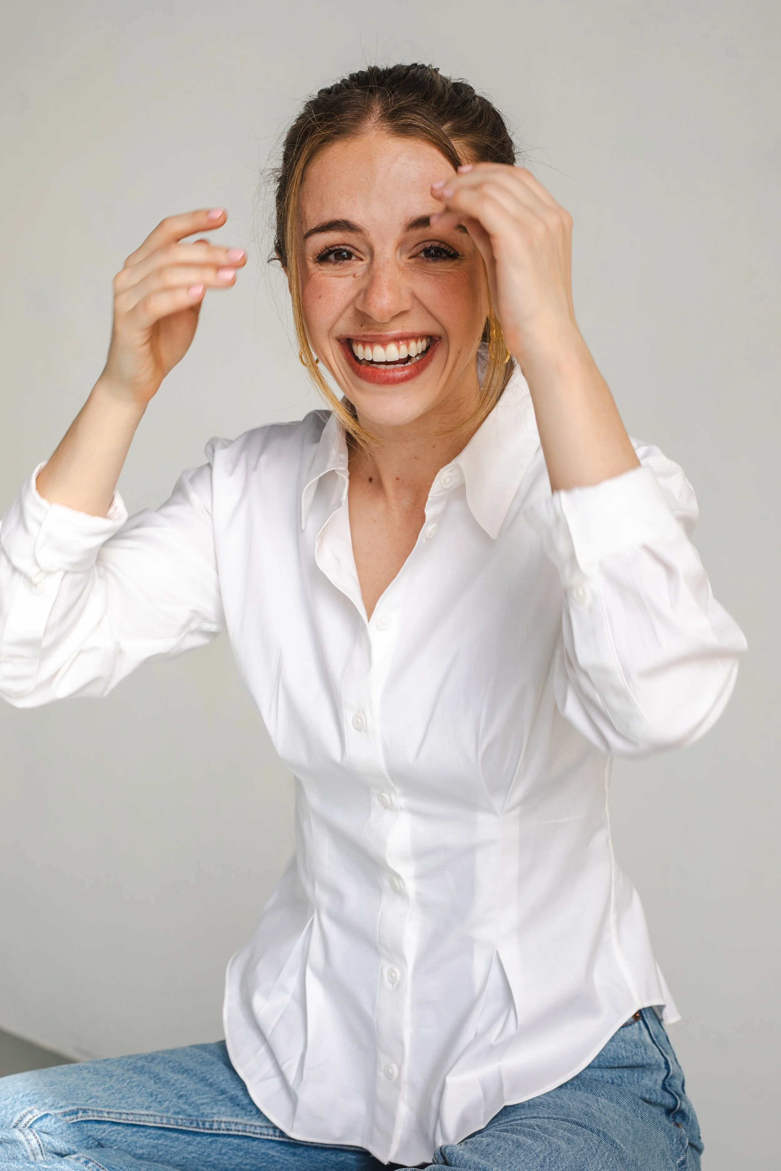 A woman with a big smile, wearing a white button-up shirt and blue jeans, sitting against a plain background.