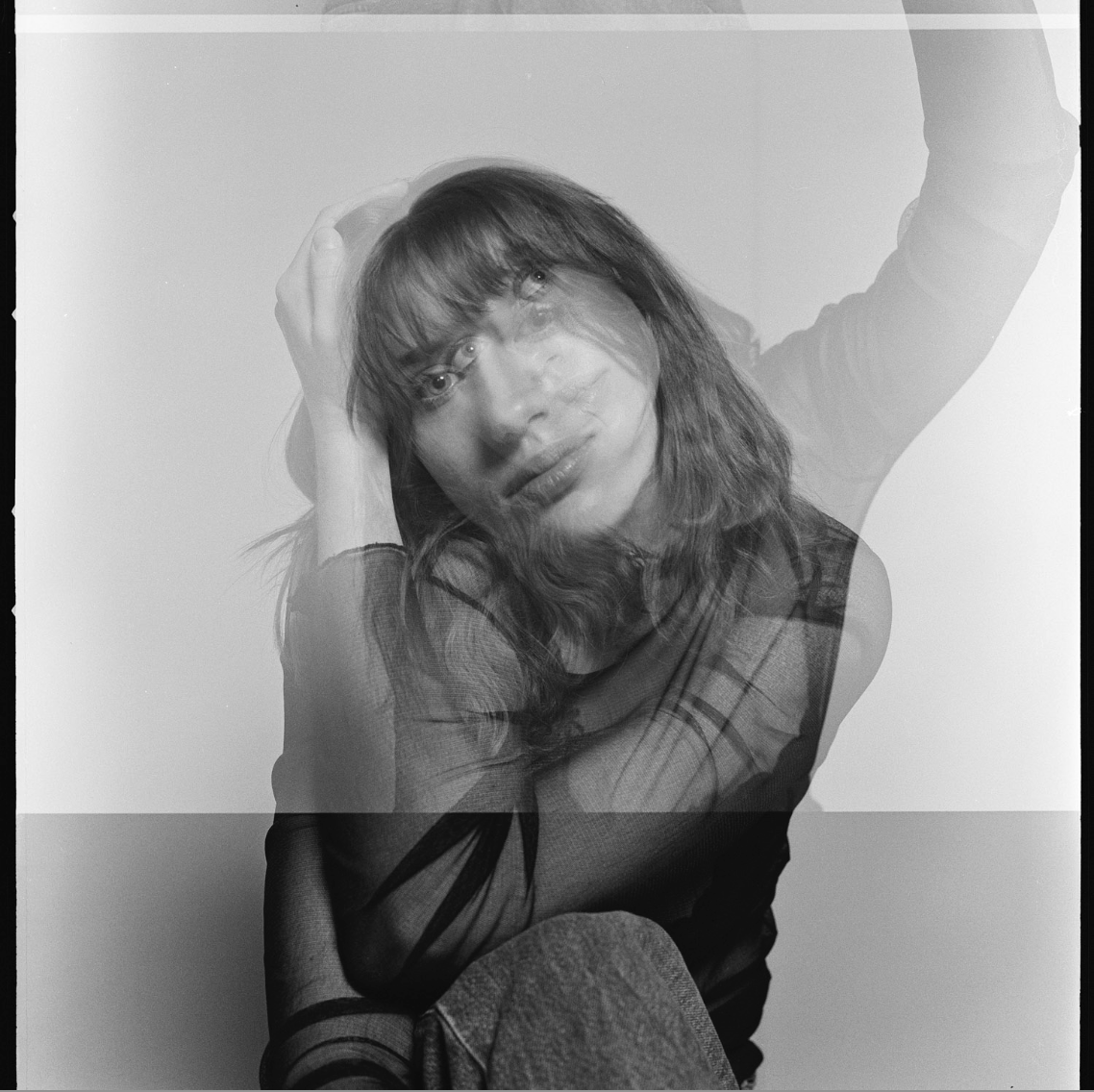 Black and white photo of a woman with shoulder-length hair, wearing a sheer top, sitting with her hand on her head, looking at the camera.