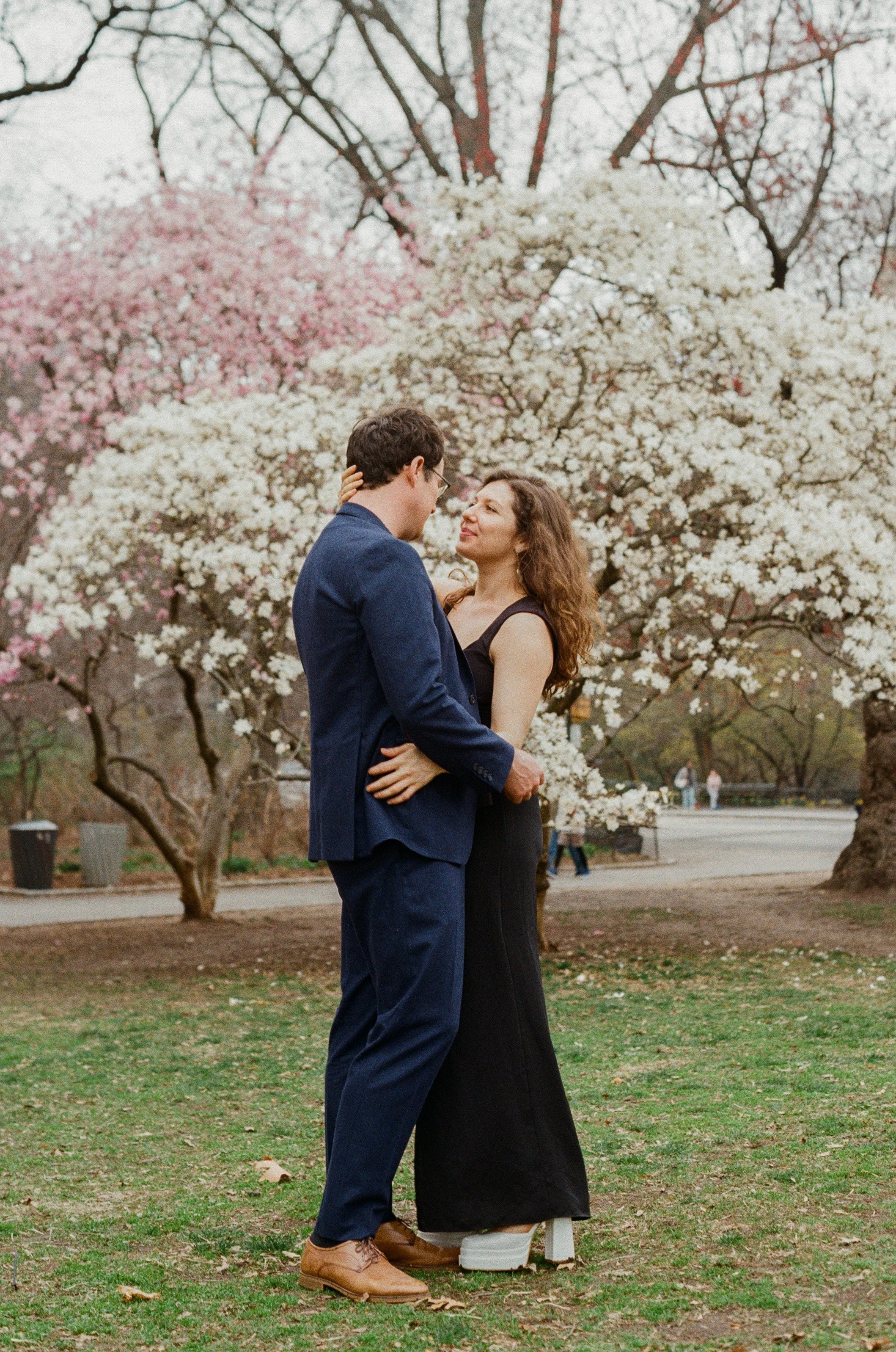 A couple standing close together in a park with blooming white and pink trees in the background.