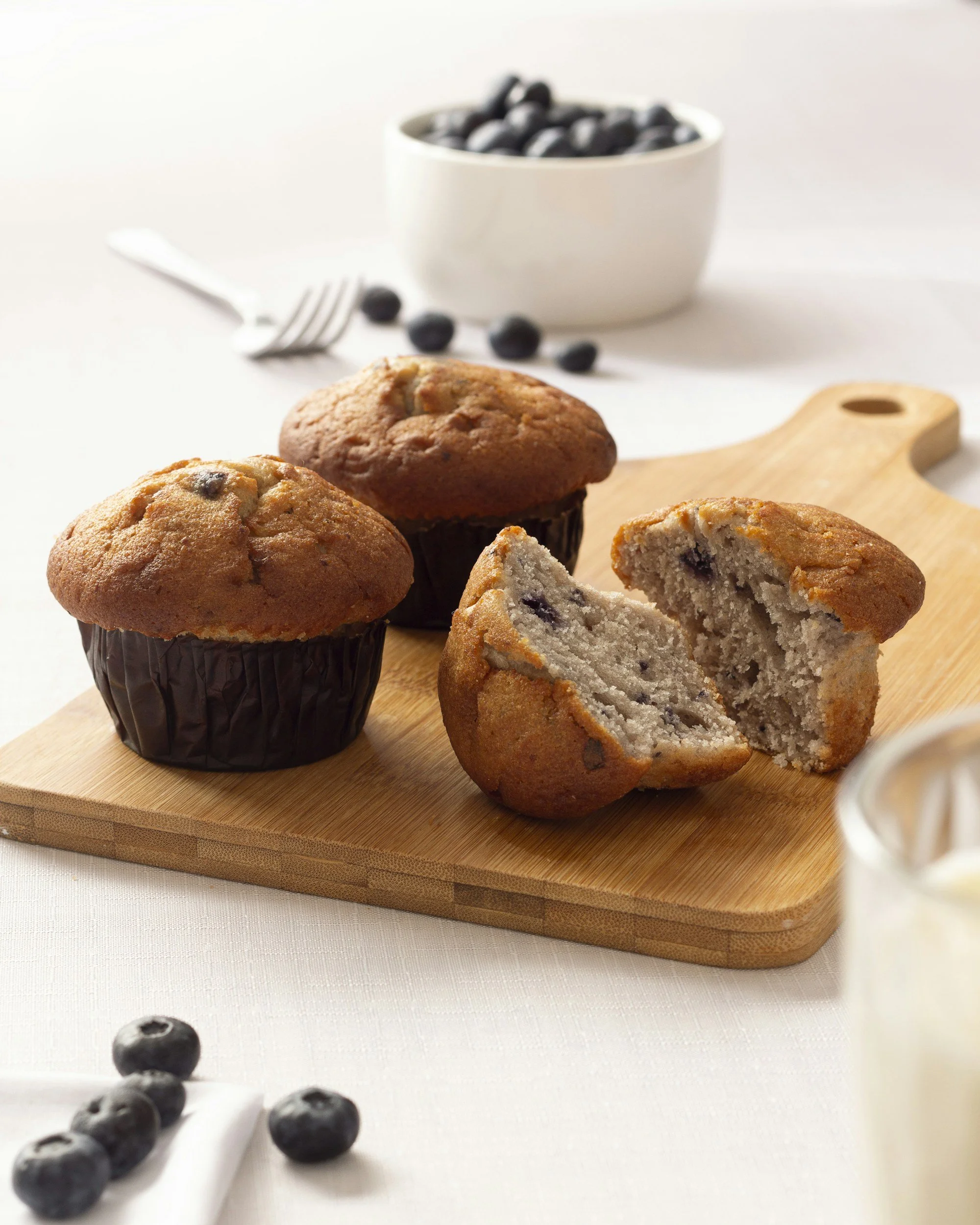 Close-up of blueberry muffins on a wooden board, with one muffin cut in half to show blueberries inside, and a bowl of blueberries and scattered blueberries behind.