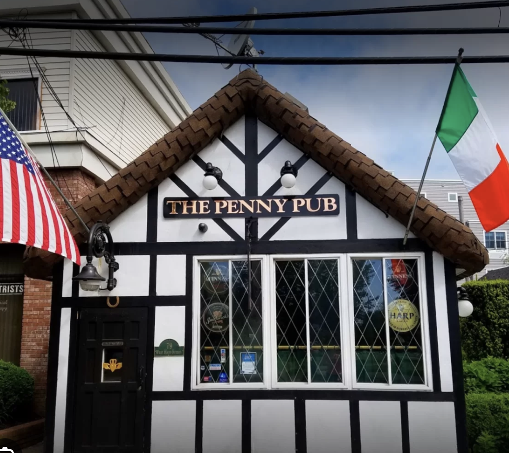 The exterior of The Penny Pub, a small building with a Tudor-style black and white facade, brown tiled roof, and windows. An American flag is on the left, and an Irish flag is on the right.