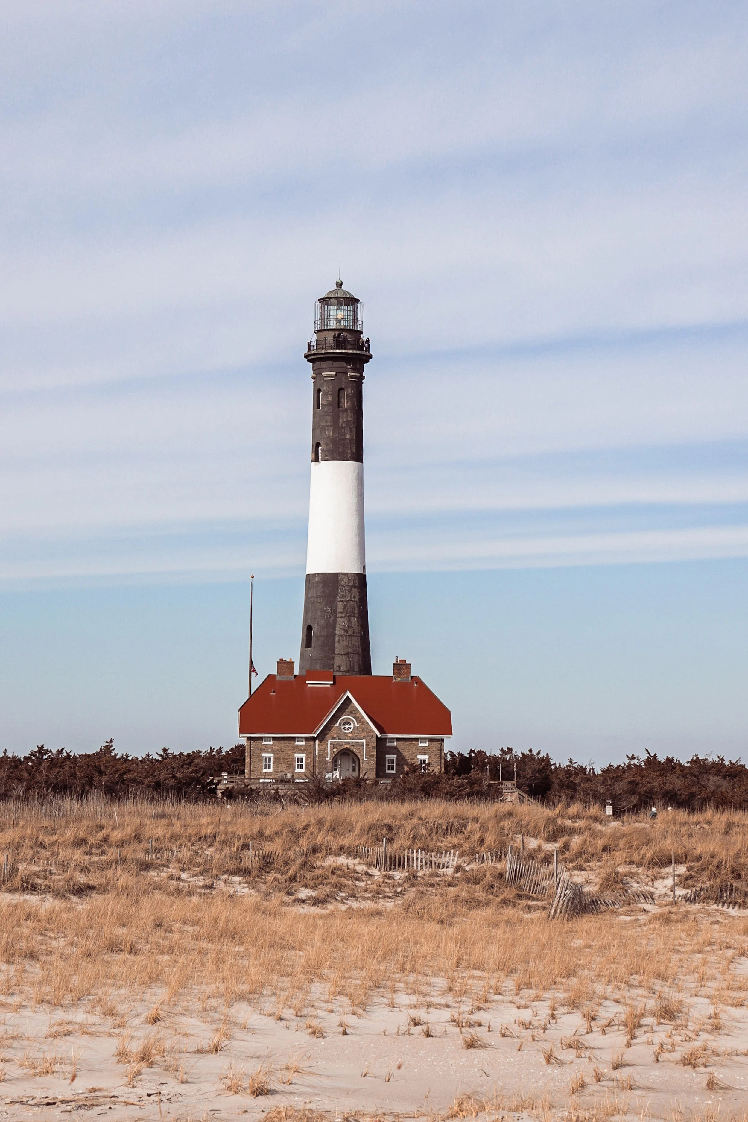 A lighthouse standing on a grassy sandy shoreline with a house at its base, under a cloudy sky.